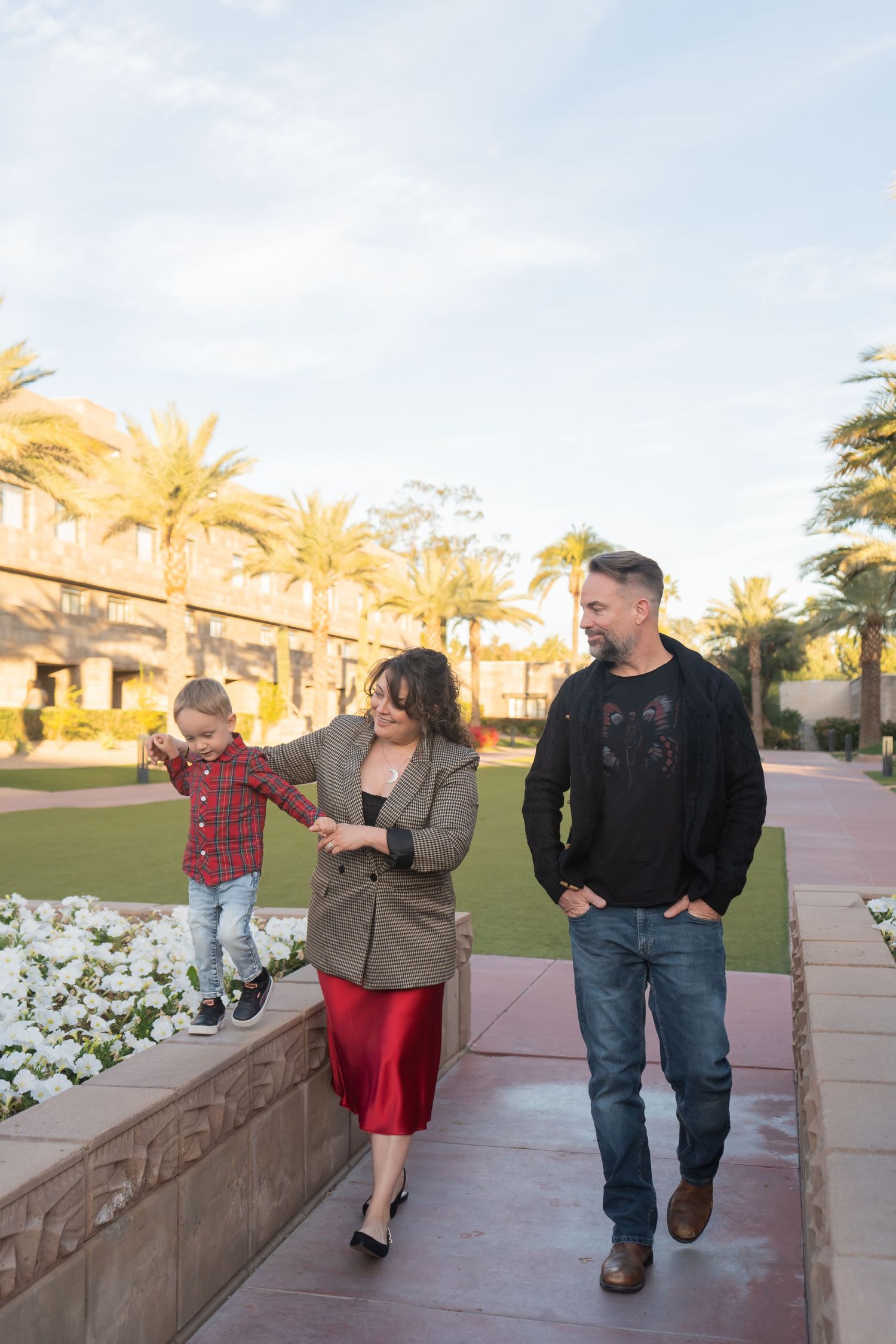 Family of three walking along garden path with palm trees, mother helping toddler balance on ledge.