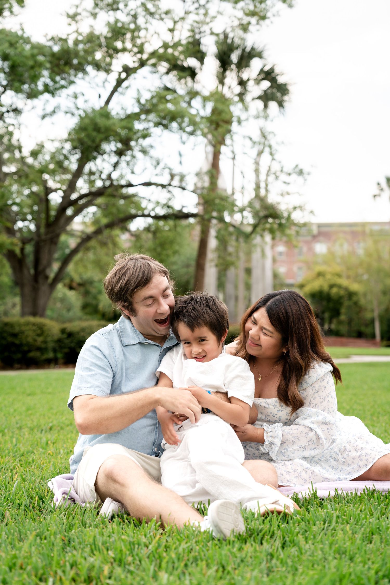A joyful family of three sits and laughs together on a grassy lawn surrounded by large shady trees.