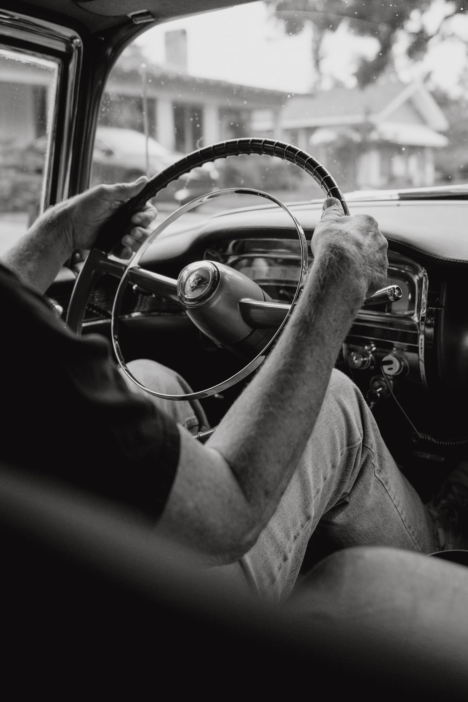 Black and white close-up of person gripping vintage car steering wheel from back seat.