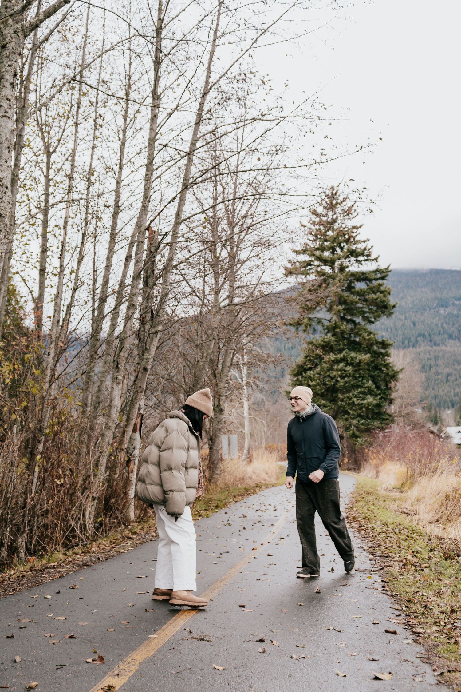 A couple in winter coats walking along a winding path lined with bare trees on an overcast day.