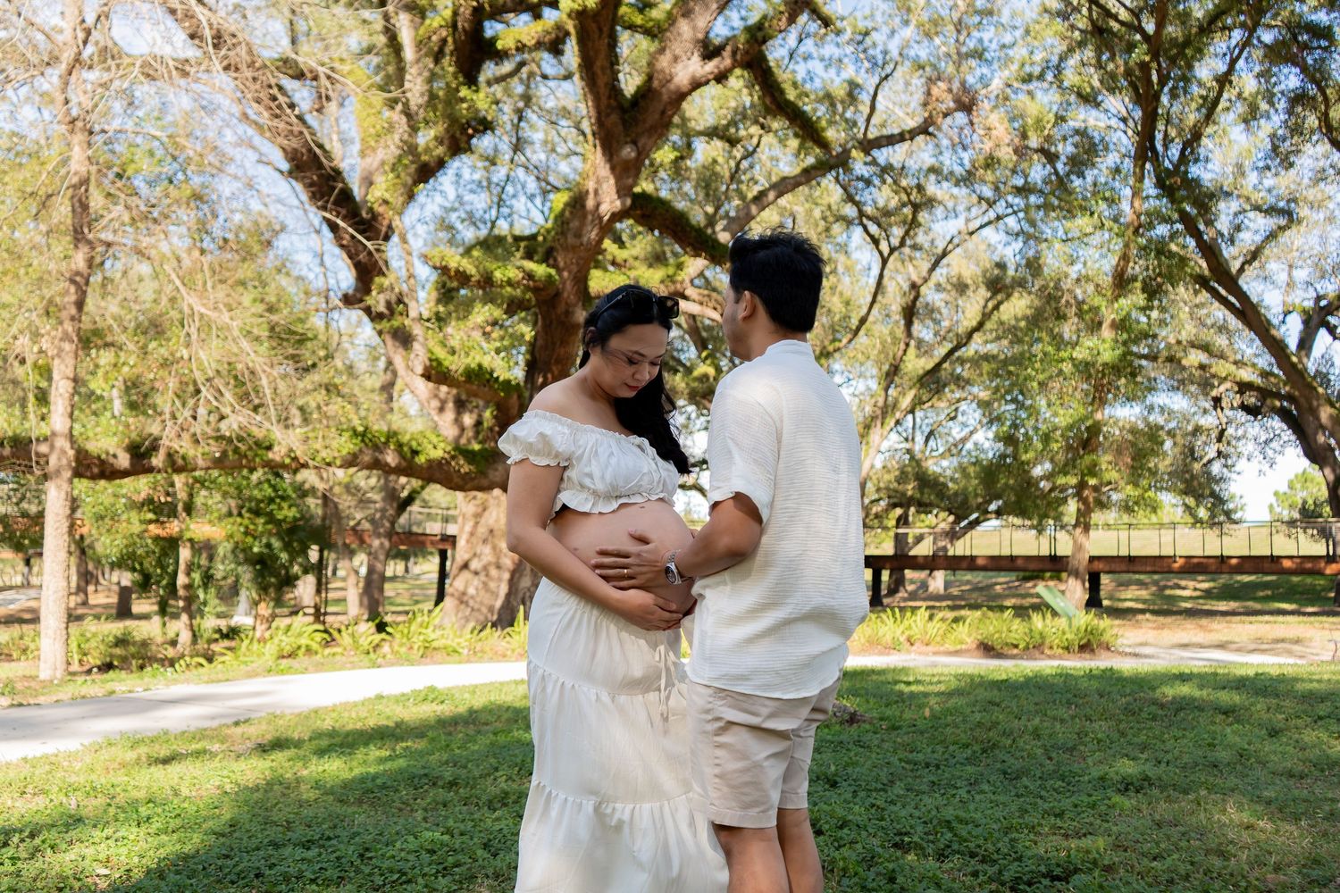 Pregnant woman in white dress with partner admiring baby bump in sunny park with large oak tree.