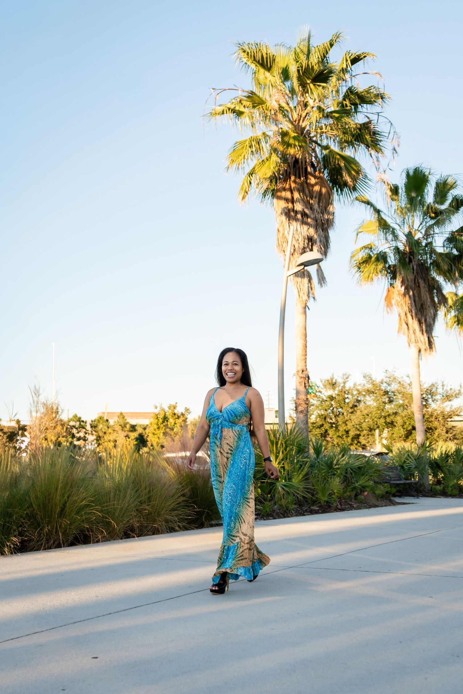Woman in blue and gold maxi dress walking on a sunny pathway with tall palm trees.