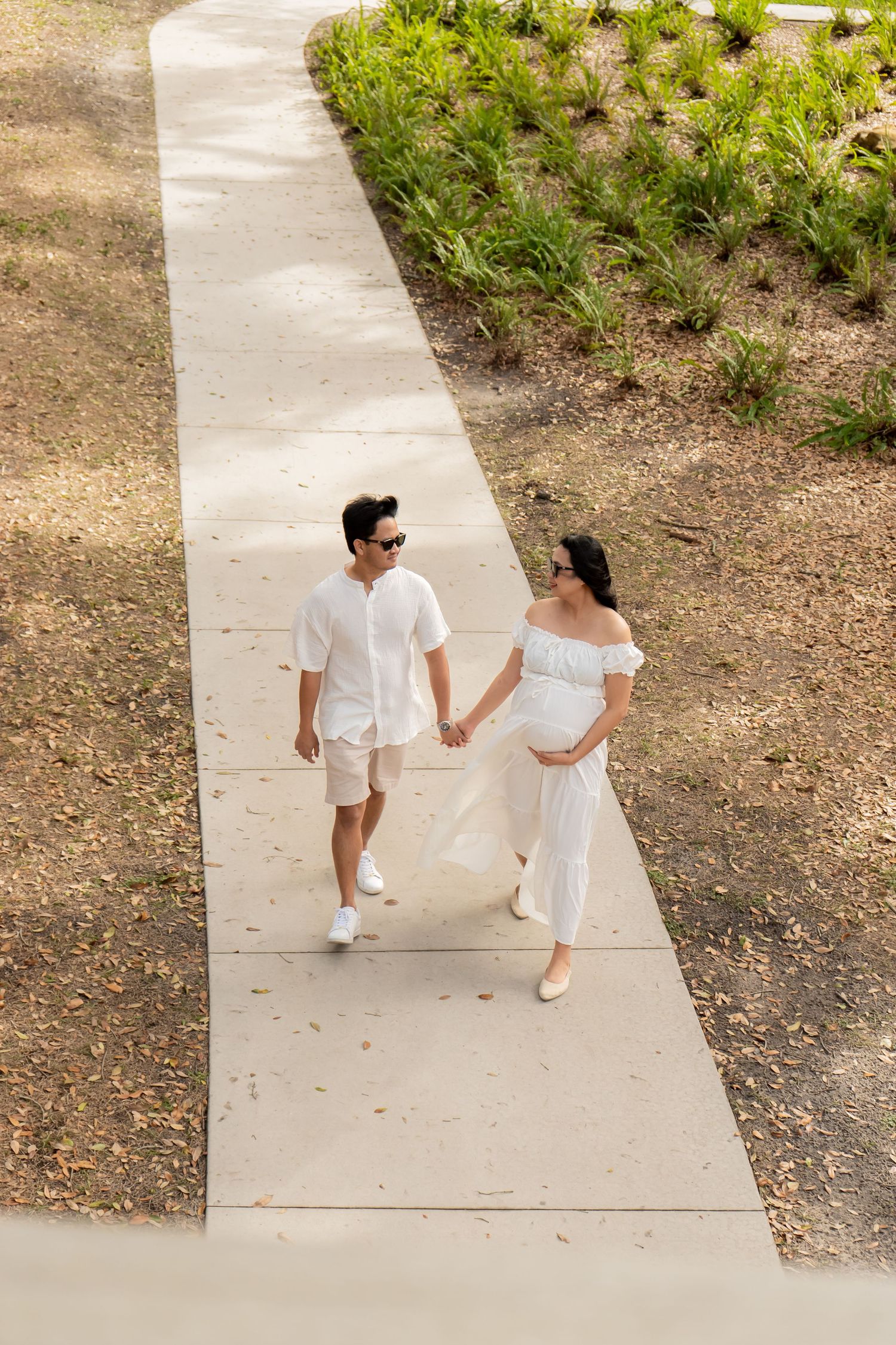 Aerial view of pregnant woman and partner walking hand in hand on sunny park pathway in white outfits.
