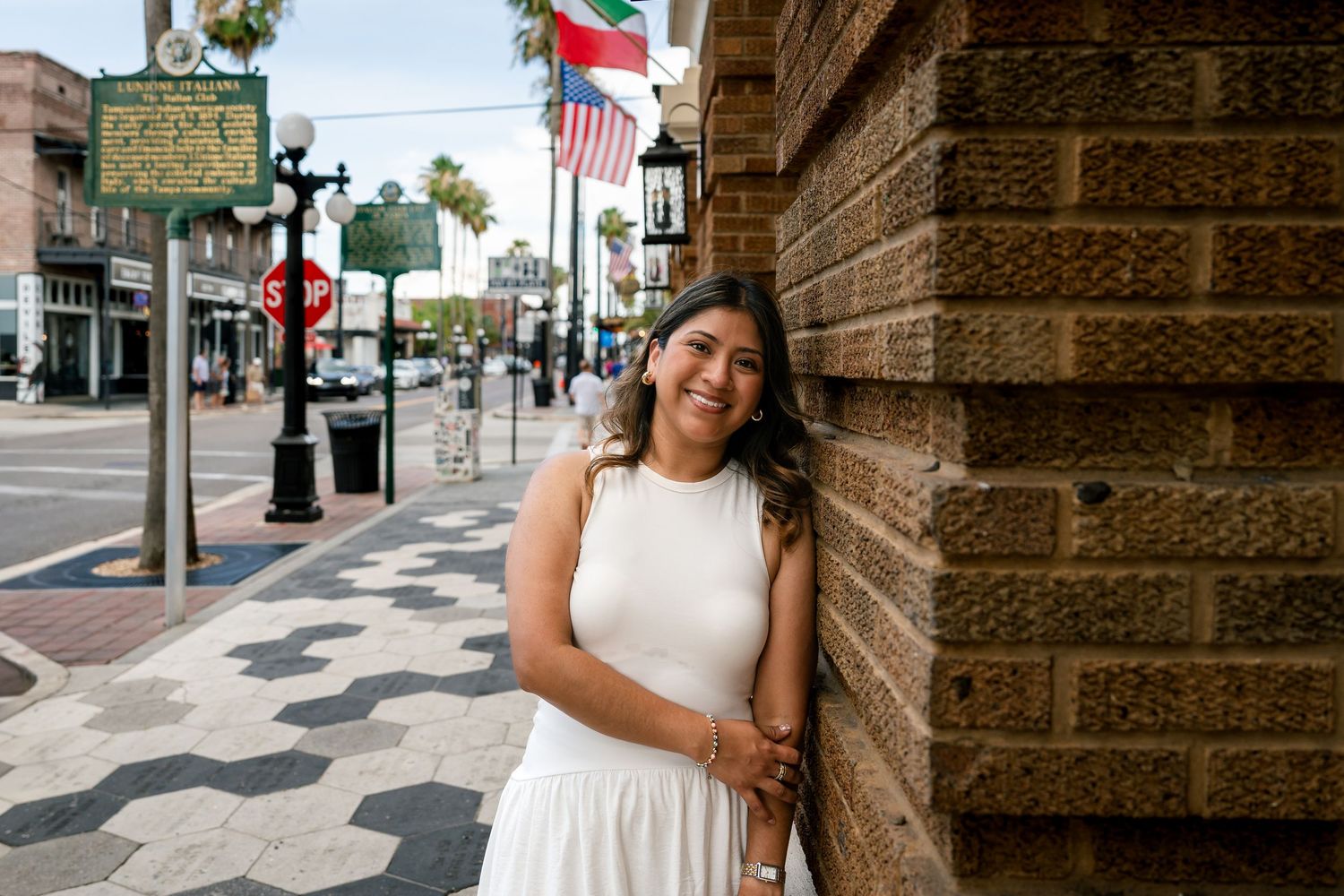 Young woman in white dress smiling and leaning against brick wall on a sunny urban street with Texas flag.