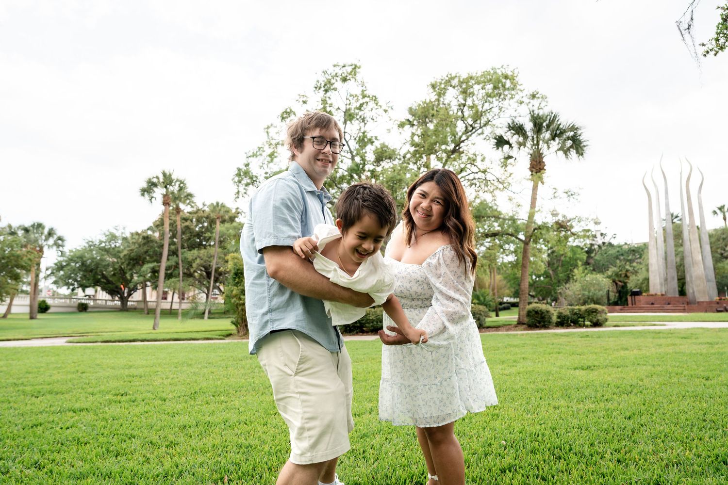 A happy family of three poses on a lush green lawn surrounded by palm trees in a Florida park.