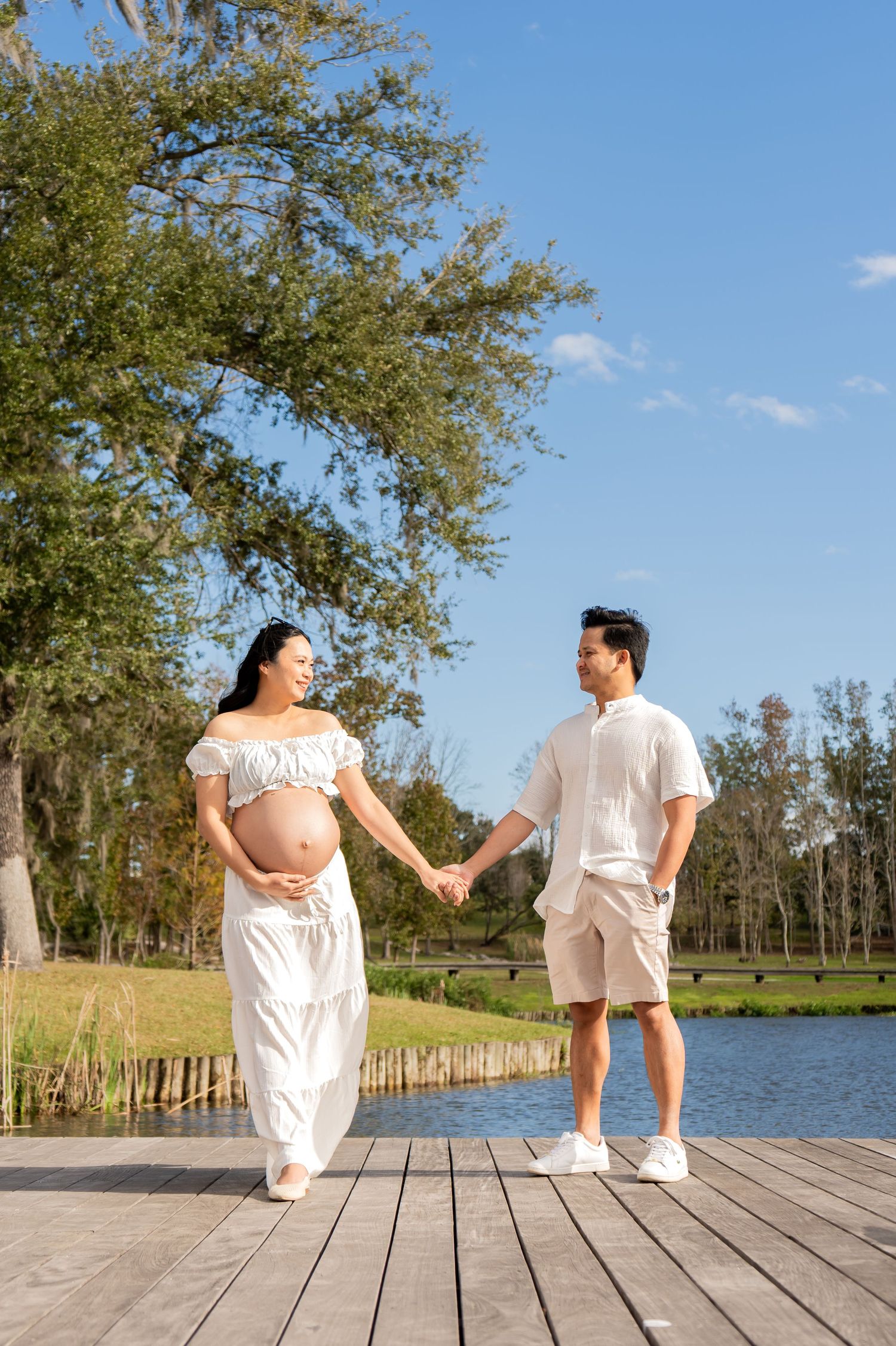 Smiling pregnant woman in white dress holds partners hand on wooden dock beside a serene lake.