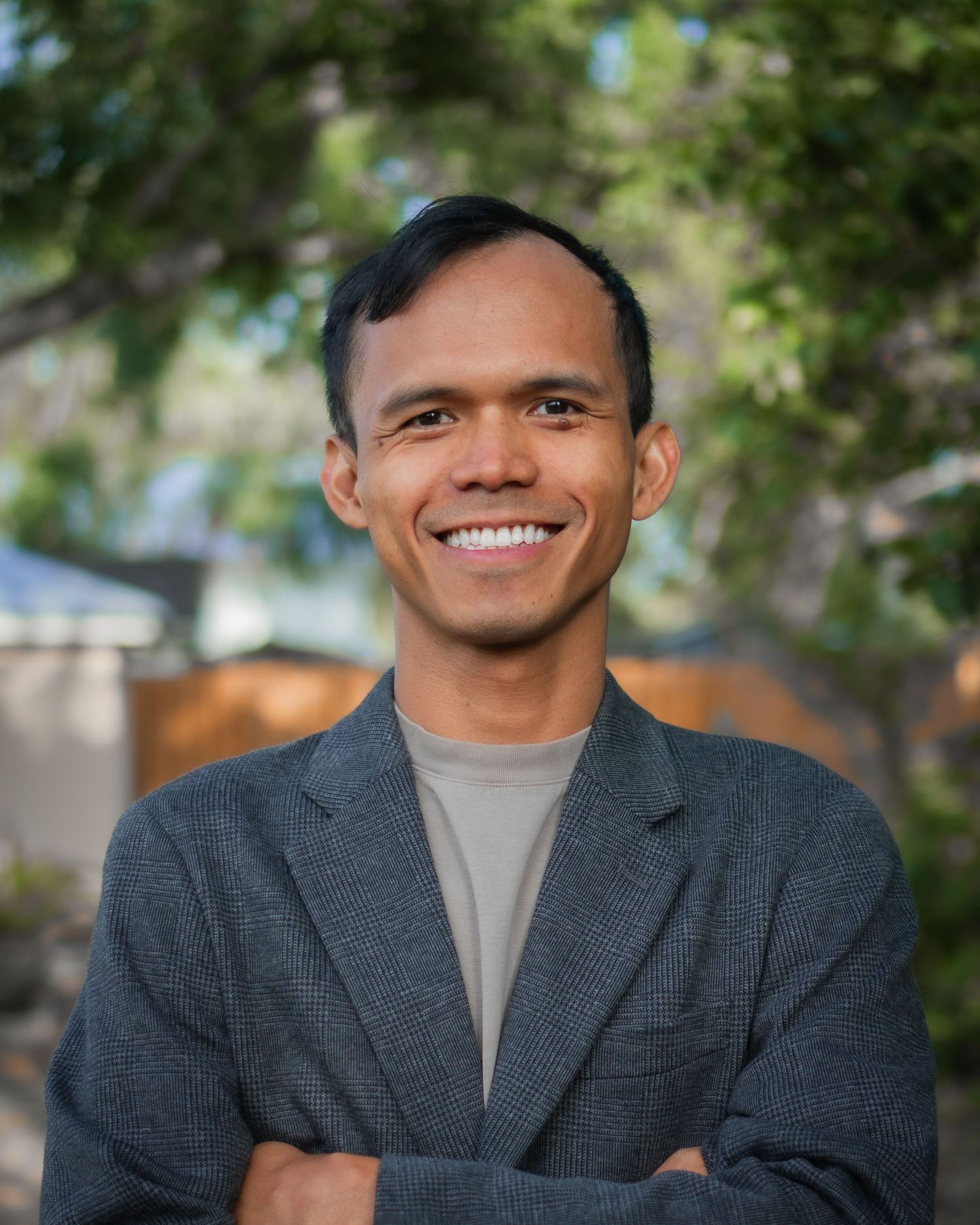 Smiling man with crossed arms wearing gray blazer poses outdoors with trees in background.