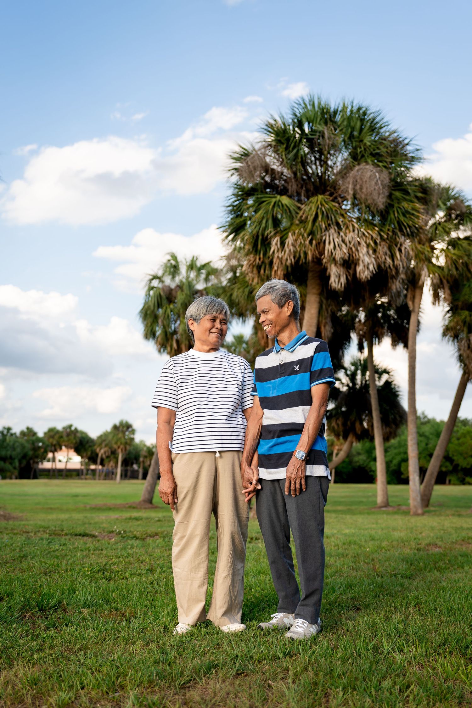 A smiling senior Asian couple holds hands in a grassy park with tall palm trees behind them.