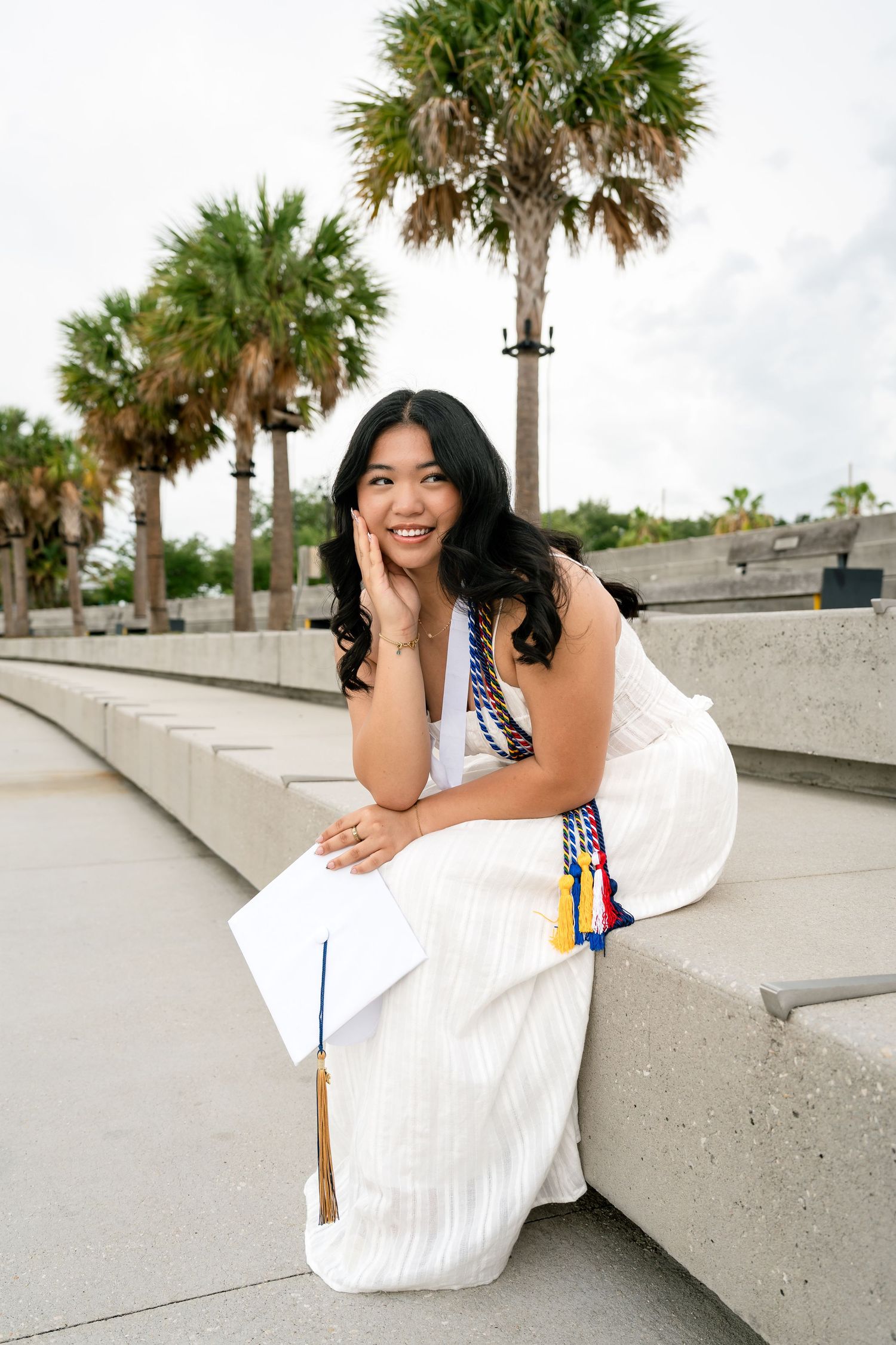 A proud smile of a graduate in a white dress holds her cap while sitting outdoors.