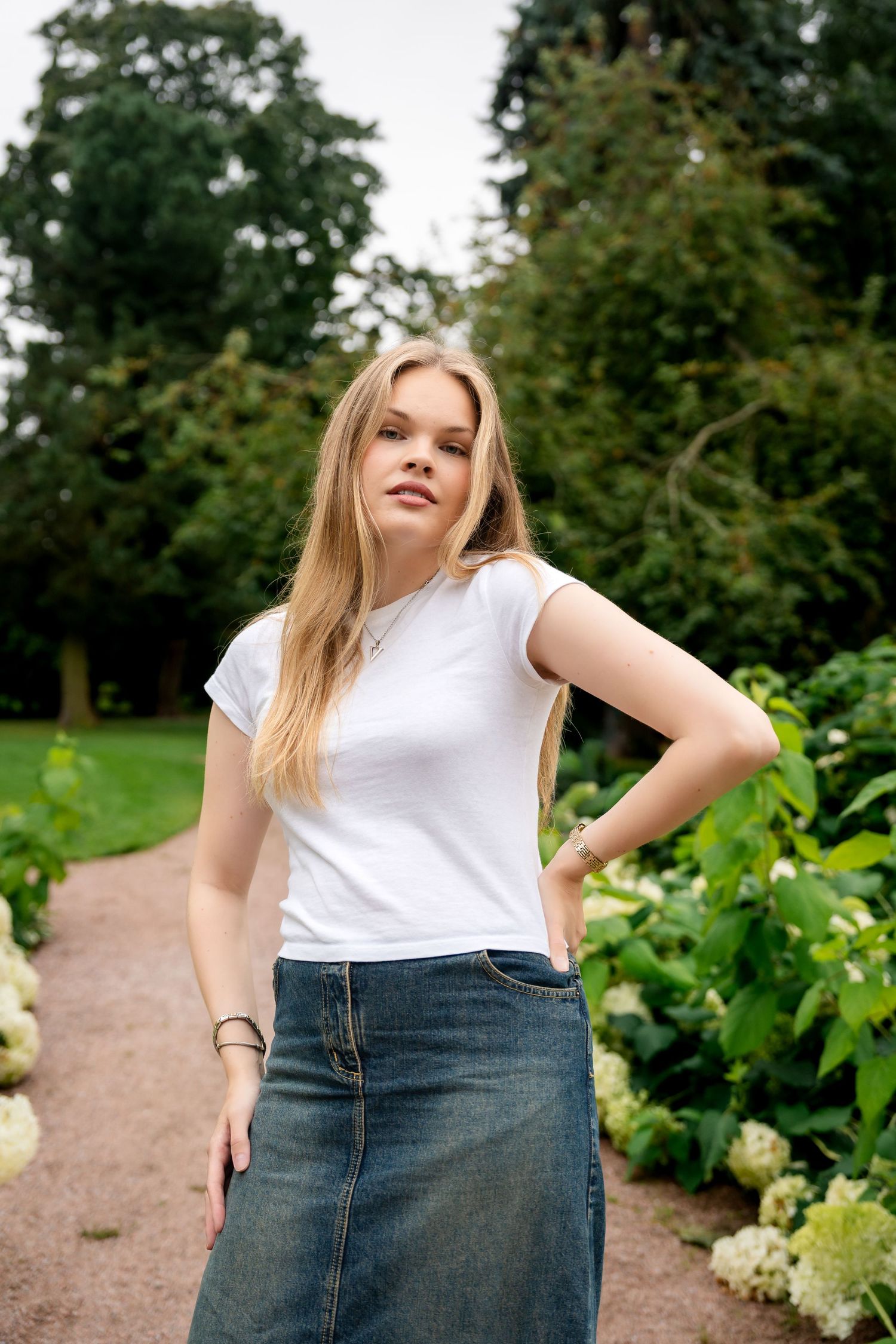 Young blonde woman in white t-shirt and denim skirt posing in a lush green park with hydrangeas.