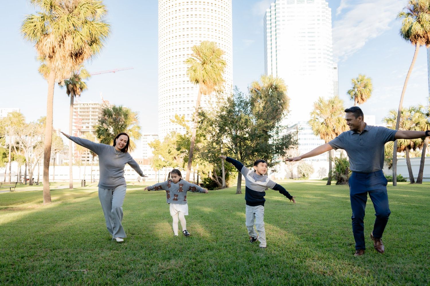 Joyful family with two children spreading arms wide in a green park with palm trees and buildings.