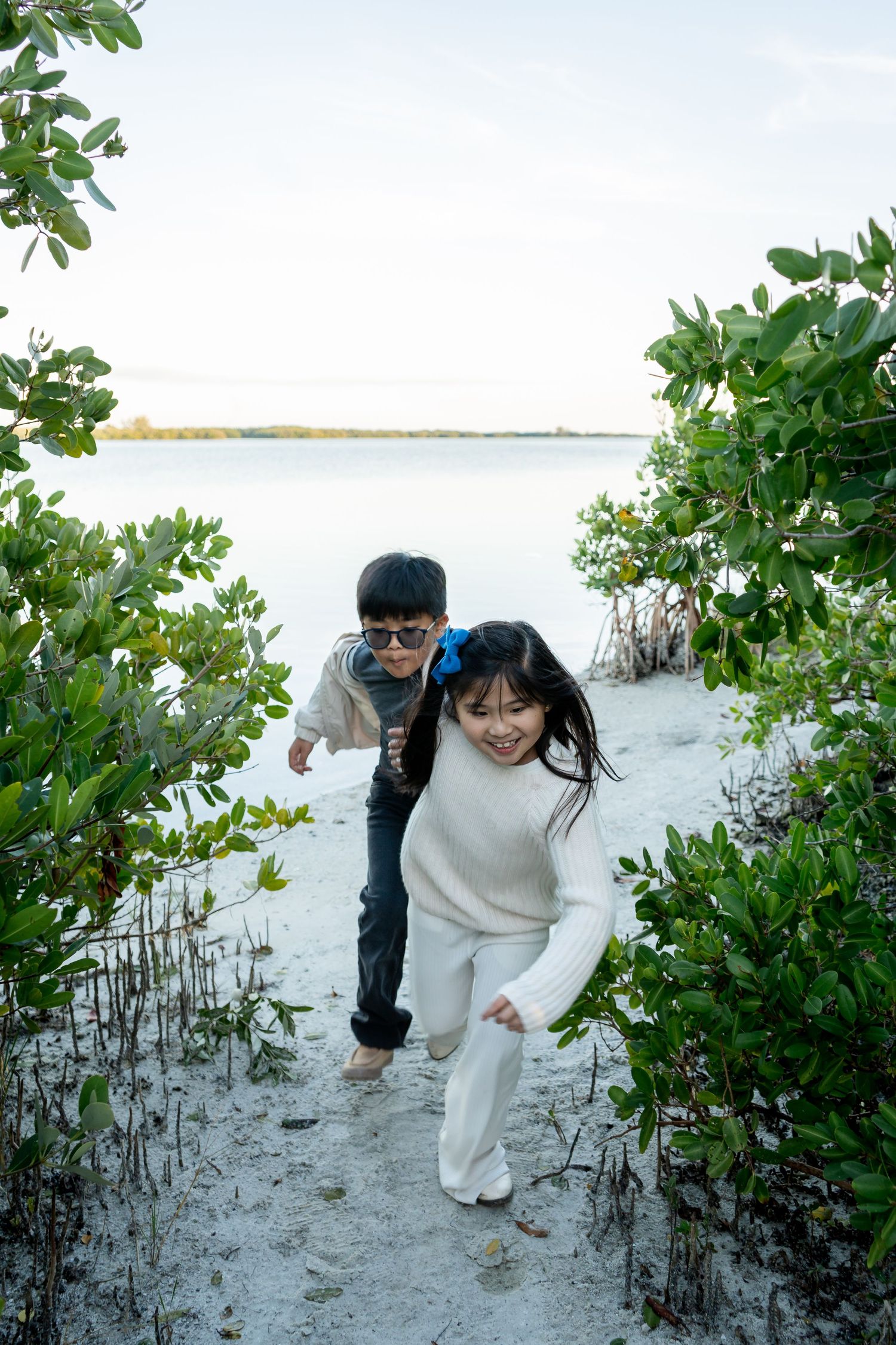 Two children running playfully through a sandy path framed by green mangrove plants near the water.