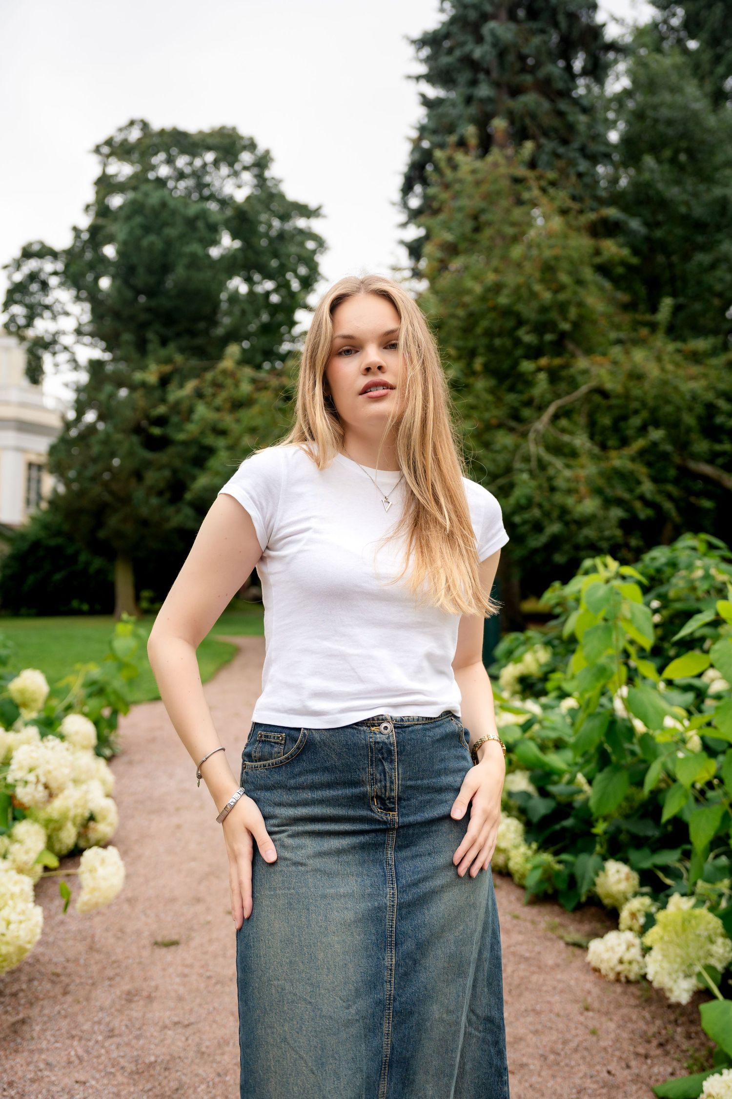 Young blonde woman in white t-shirt and denim maxi skirt posing in a lush garden path with hydrangeas.