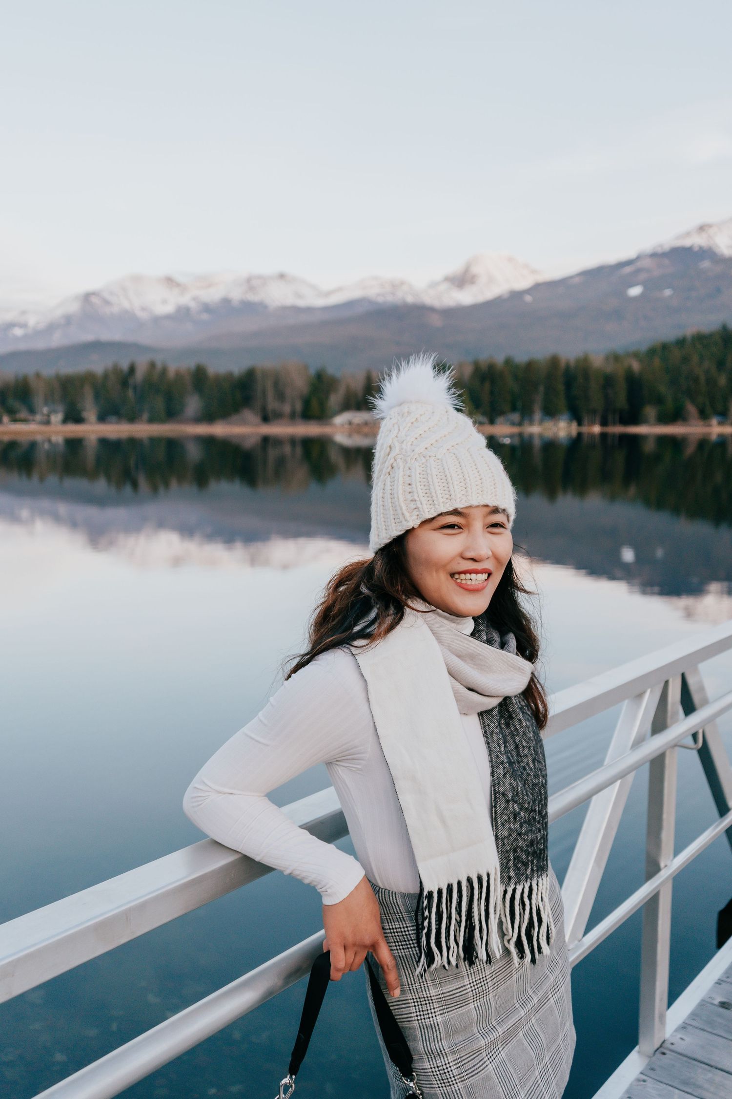 Smiling woman in knit hat and scarf leans on railing with snowy mountain lake reflection.