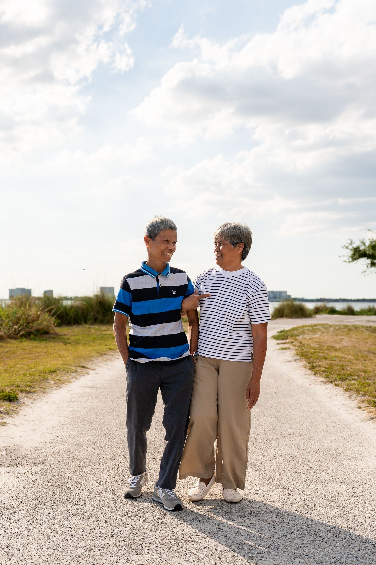 A senior Asian couple walks hand in hand along a scenic waterfront path under a cloudy sky.