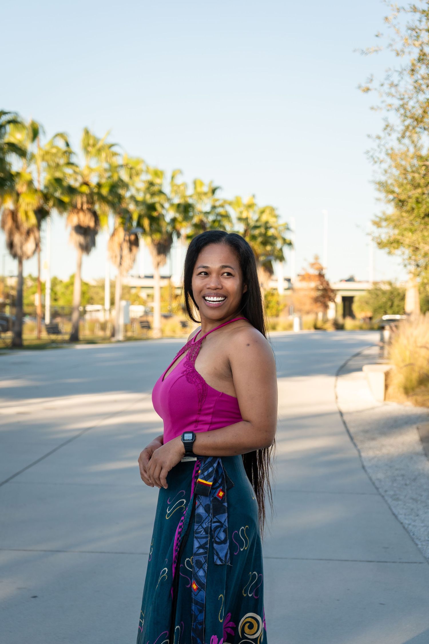 Happy woman in pink halter top and teal patterned skirt smiling on an outdoor pathway.
