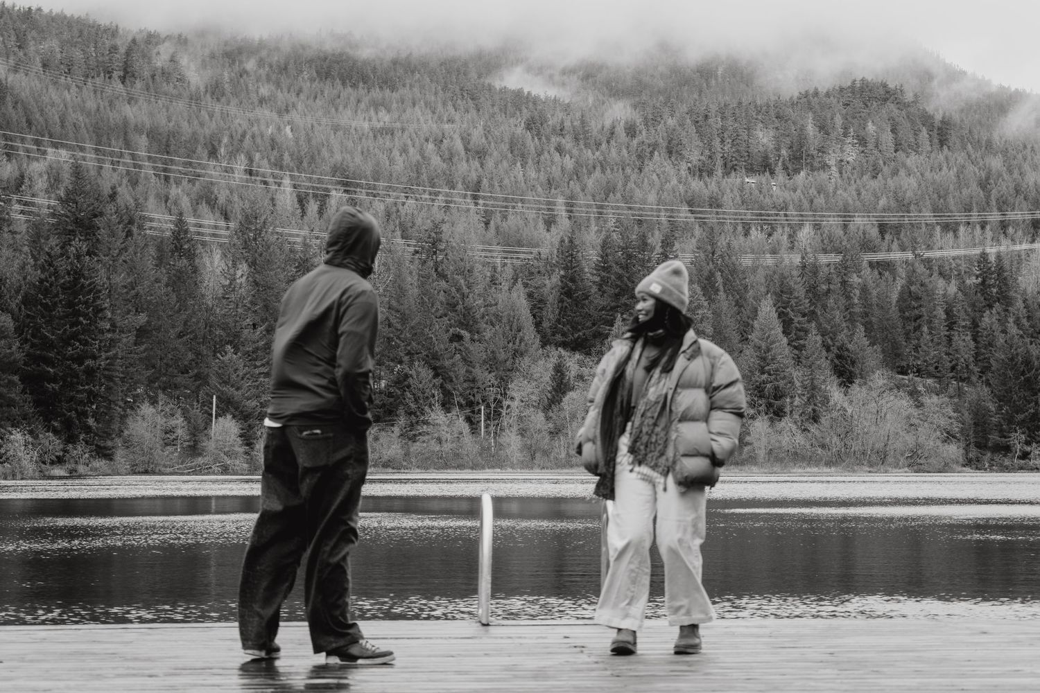 Two people in hooded jackets talking on dock by misty mountain lake in black and white.