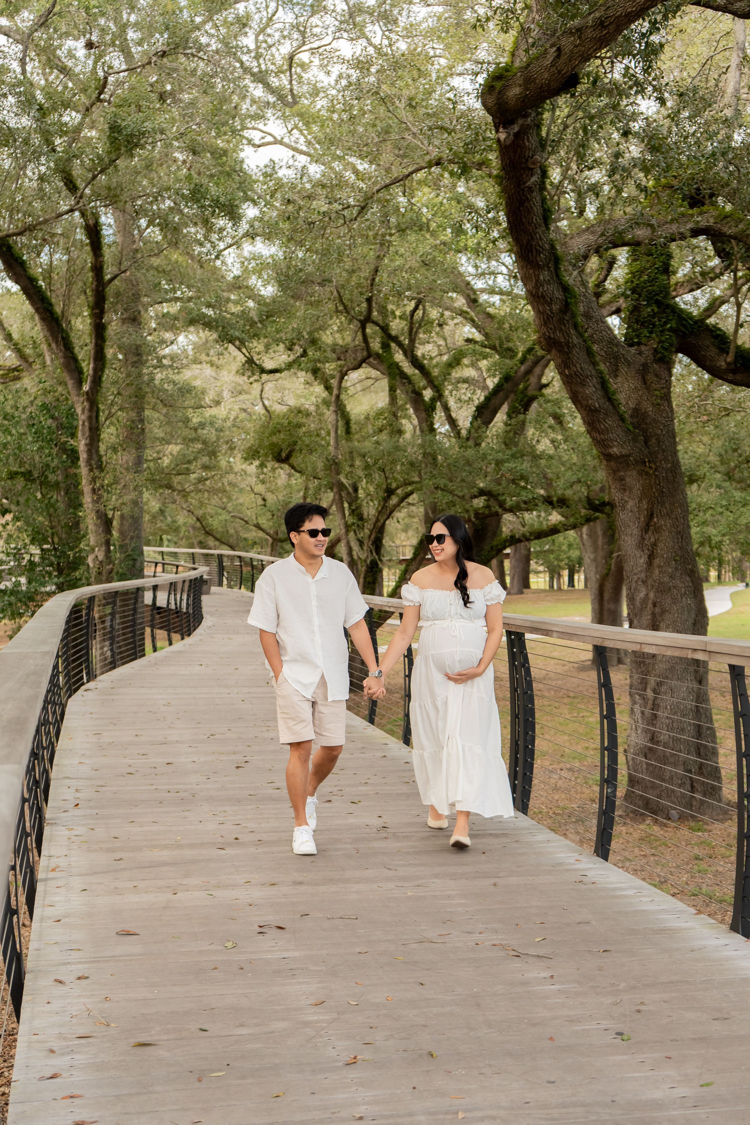 Expecting couple in white outfits holding hands walking on a wooden bridge surrounded by lush green trees.