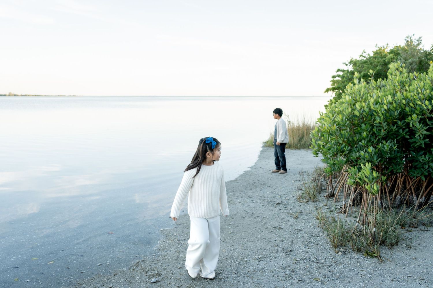 A girl in white stands near calm water while a boy looks toward lush green mangroves on a shoreline.