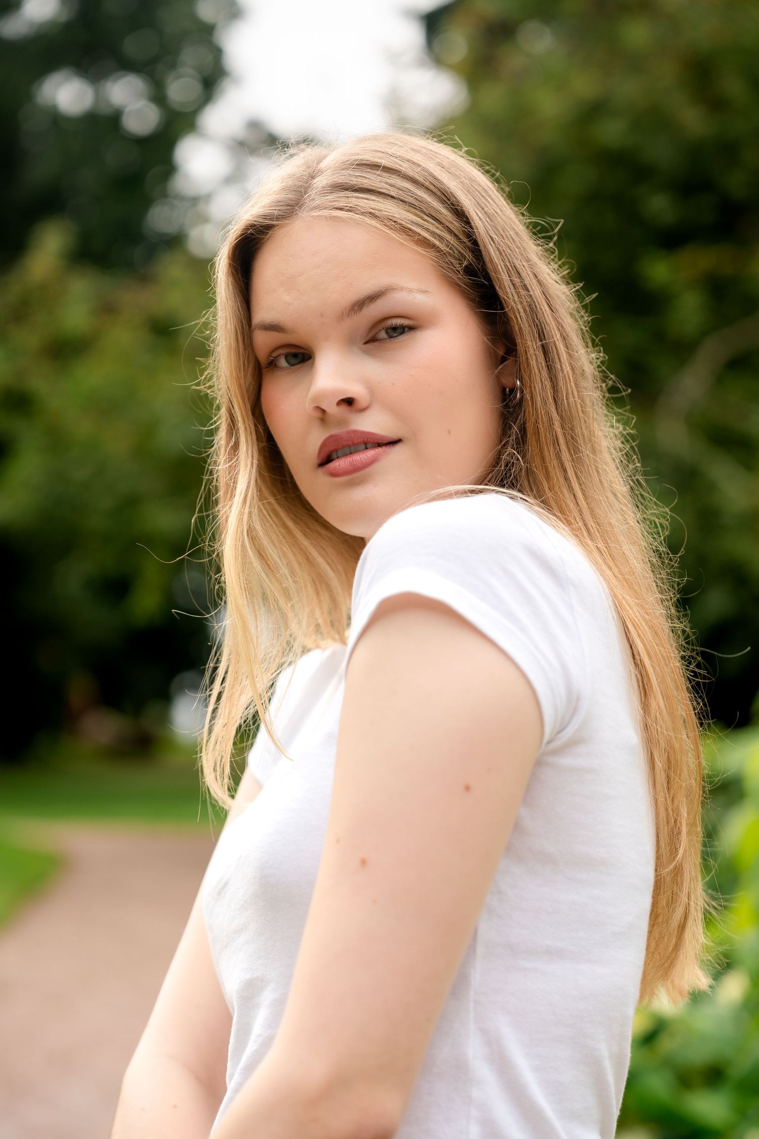 Young blonde woman in white t-shirt poses outdoors in a lush green park, looking over her shoulder.