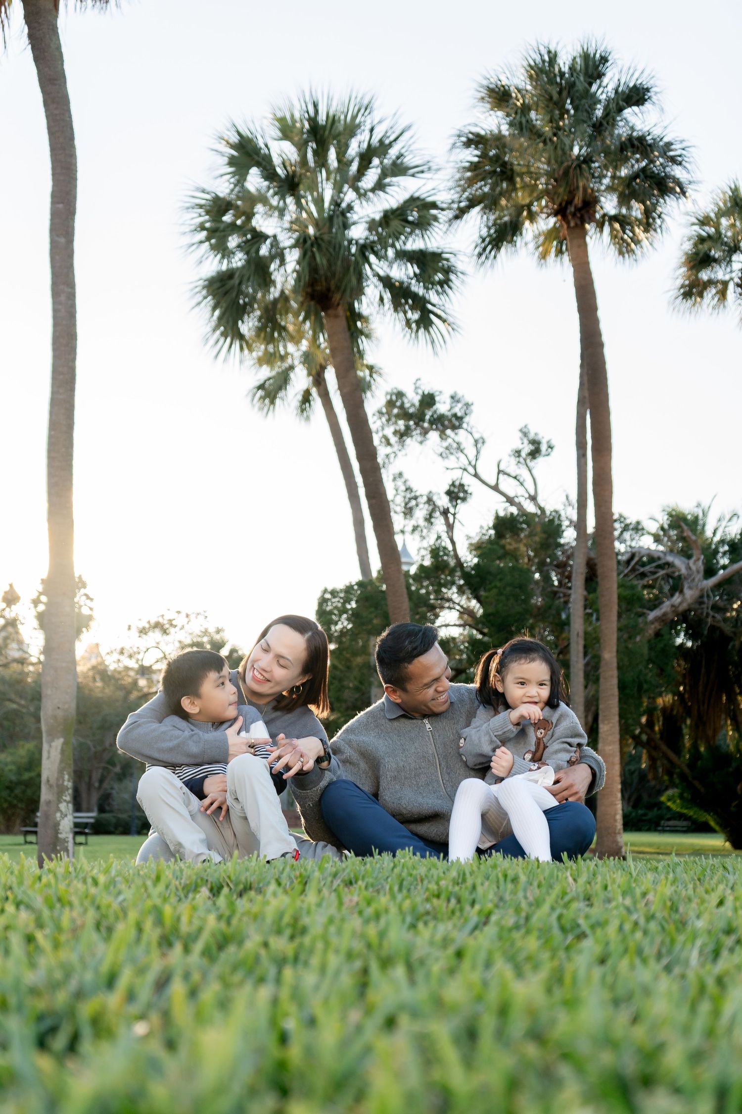 Happy family of four sitting together on grass in a park with tall palm trees at sunset.
