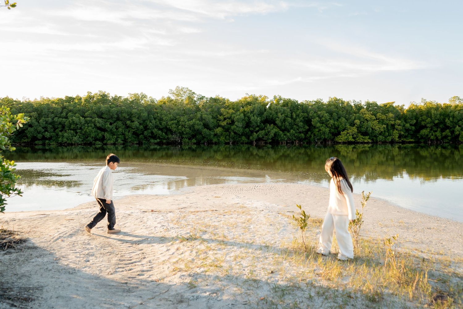 Two children walk along a sandy shoreline beside calm water with lush green mangroves in the background.