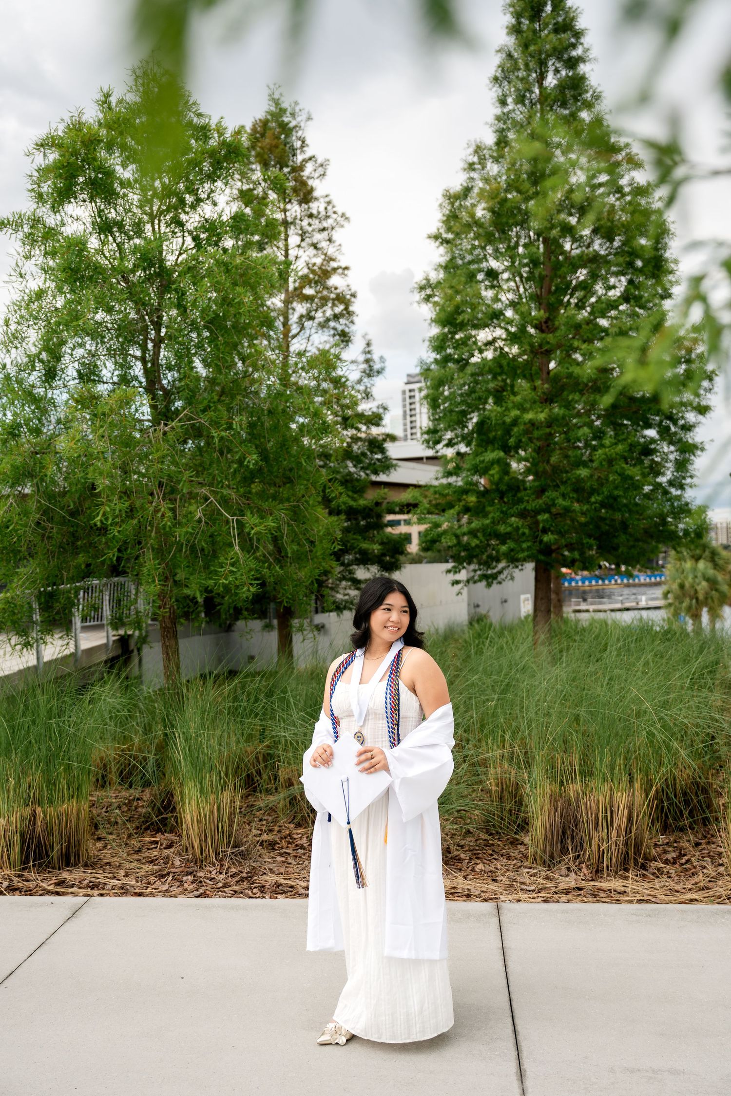 Young woman in white graduation gown and stole poses outdoors among lush green trees and ornamental grasses.