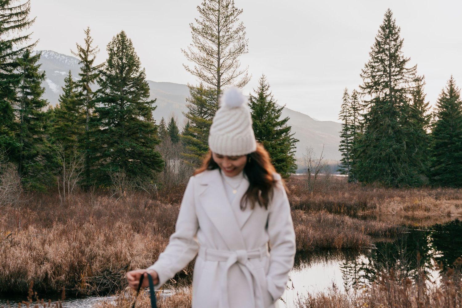 Woman in white belted coat and pom beanie walks near pond with evergreen trees.