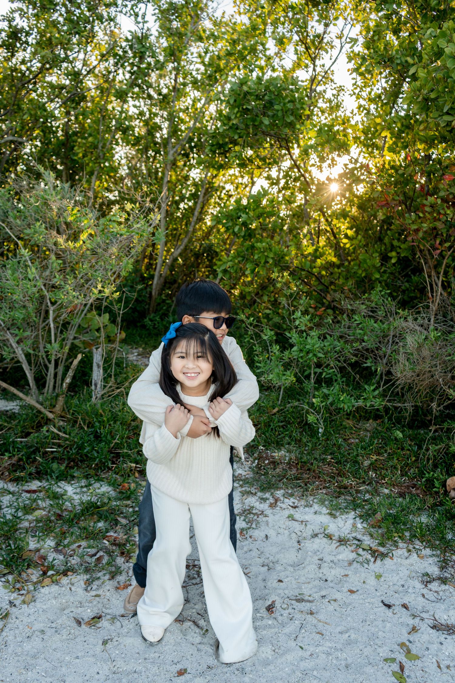A young girl smiles while being hugged from behind by another child in a lush green outdoor setting.