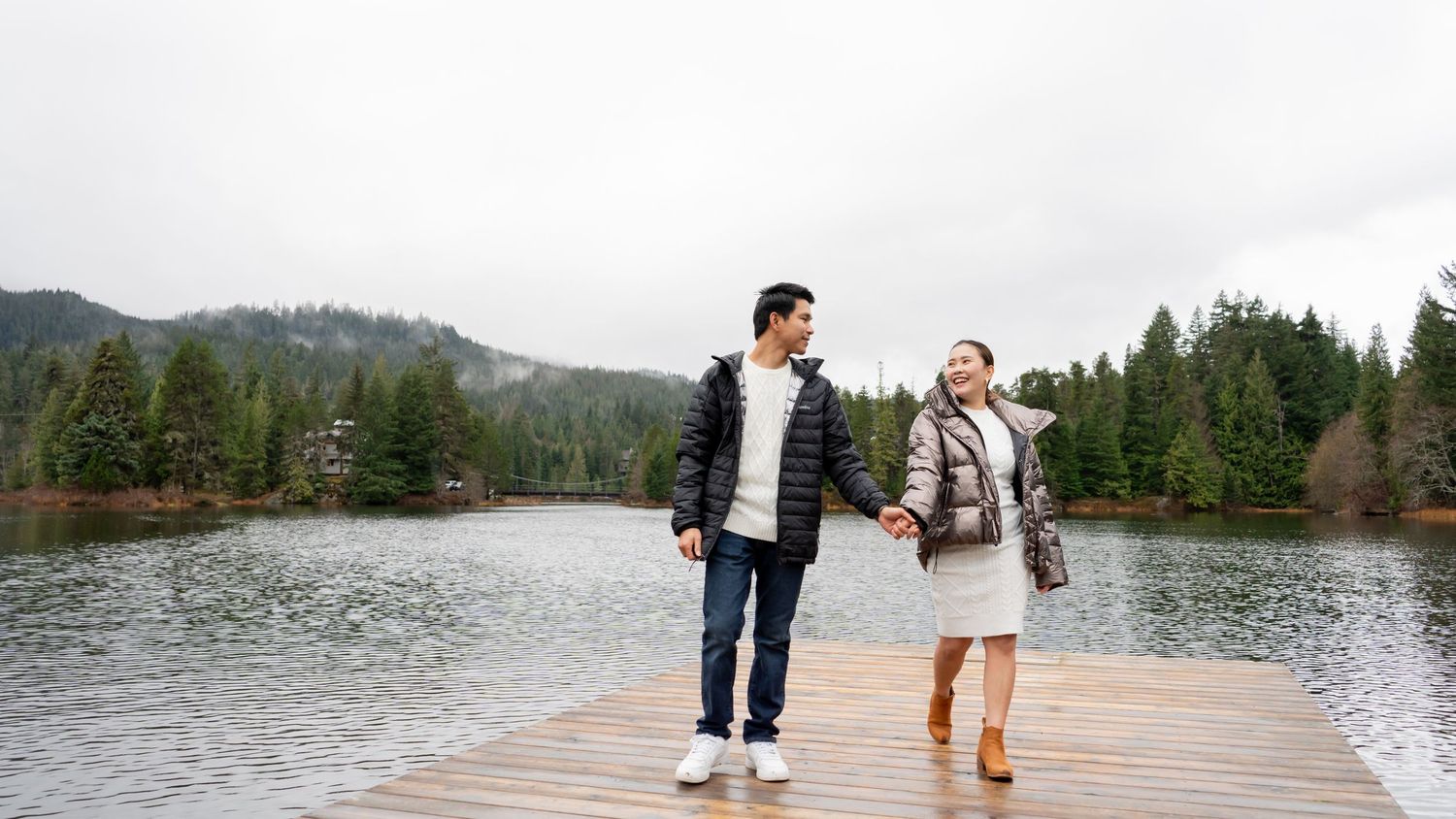 Couple holding hands walking on wooden dock with serene lake and forest backdrop.
