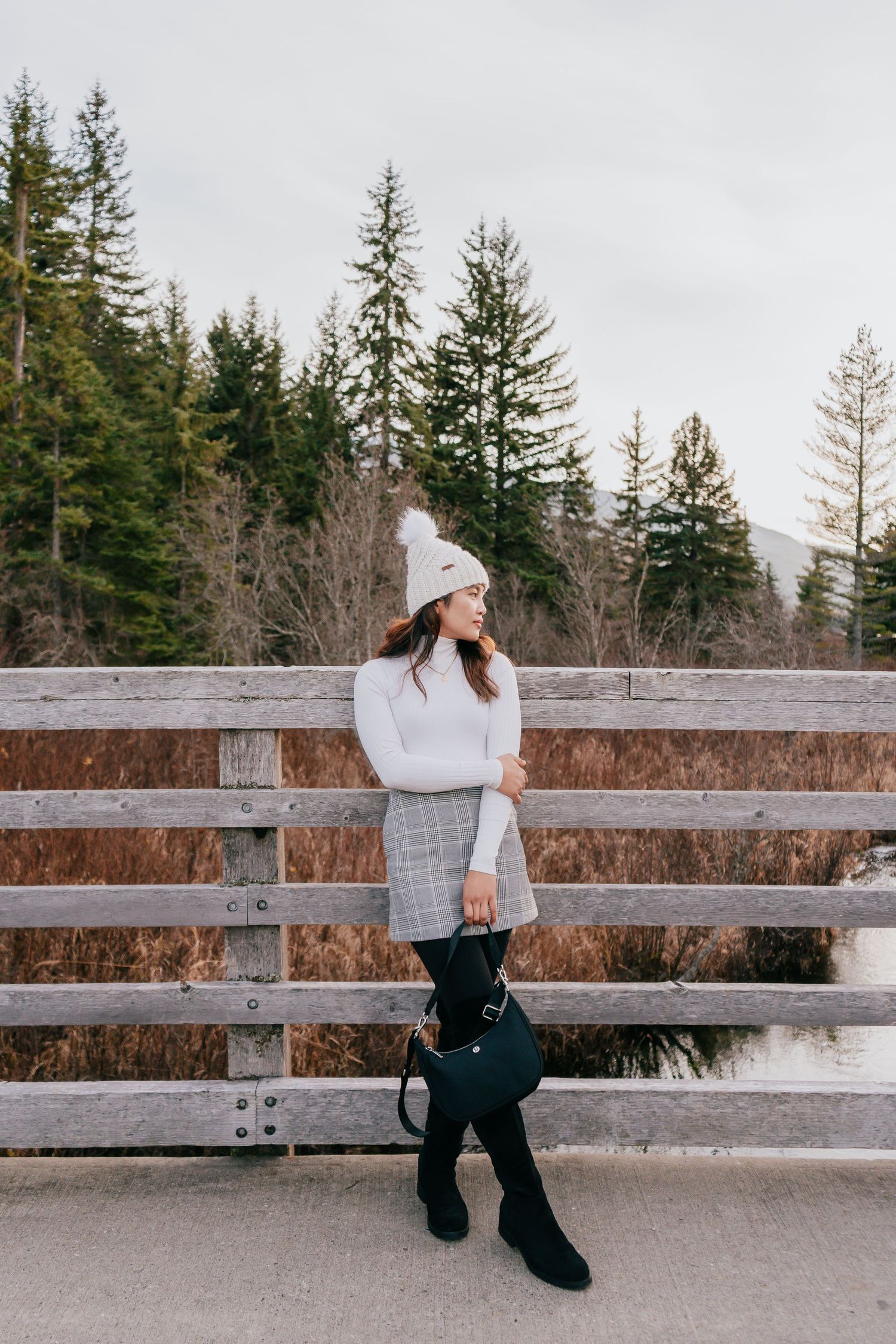 Woman in plaid skirt and white knit hat poses against wooden bridge railing with pine trees.