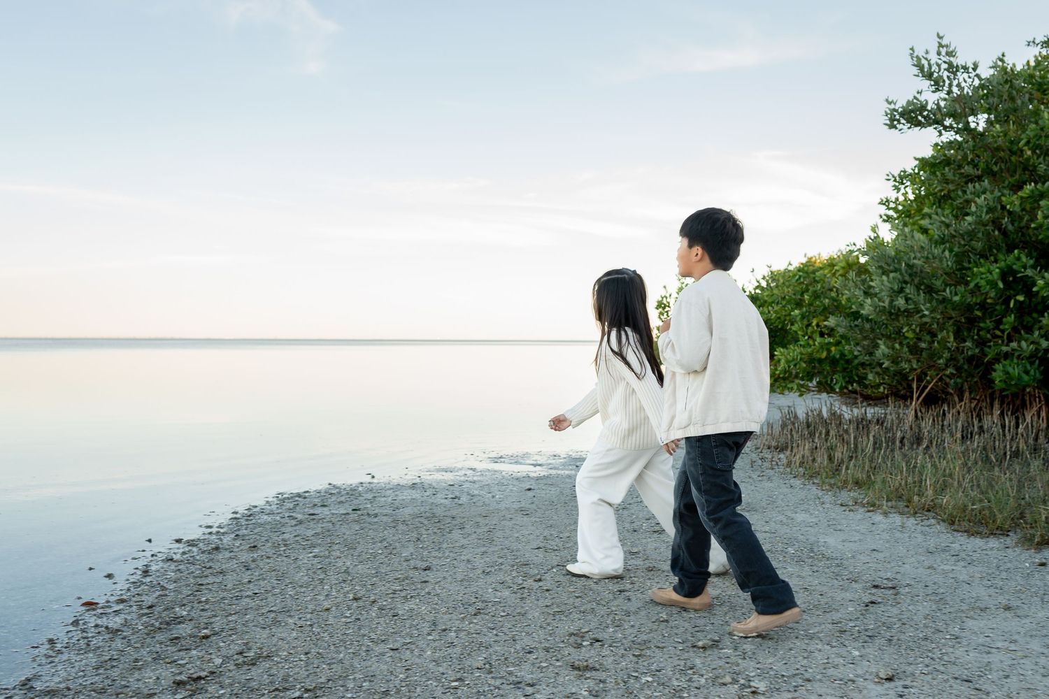 Two children in white and dark outfits walk hand in hand along a rocky shoreline at dusk.