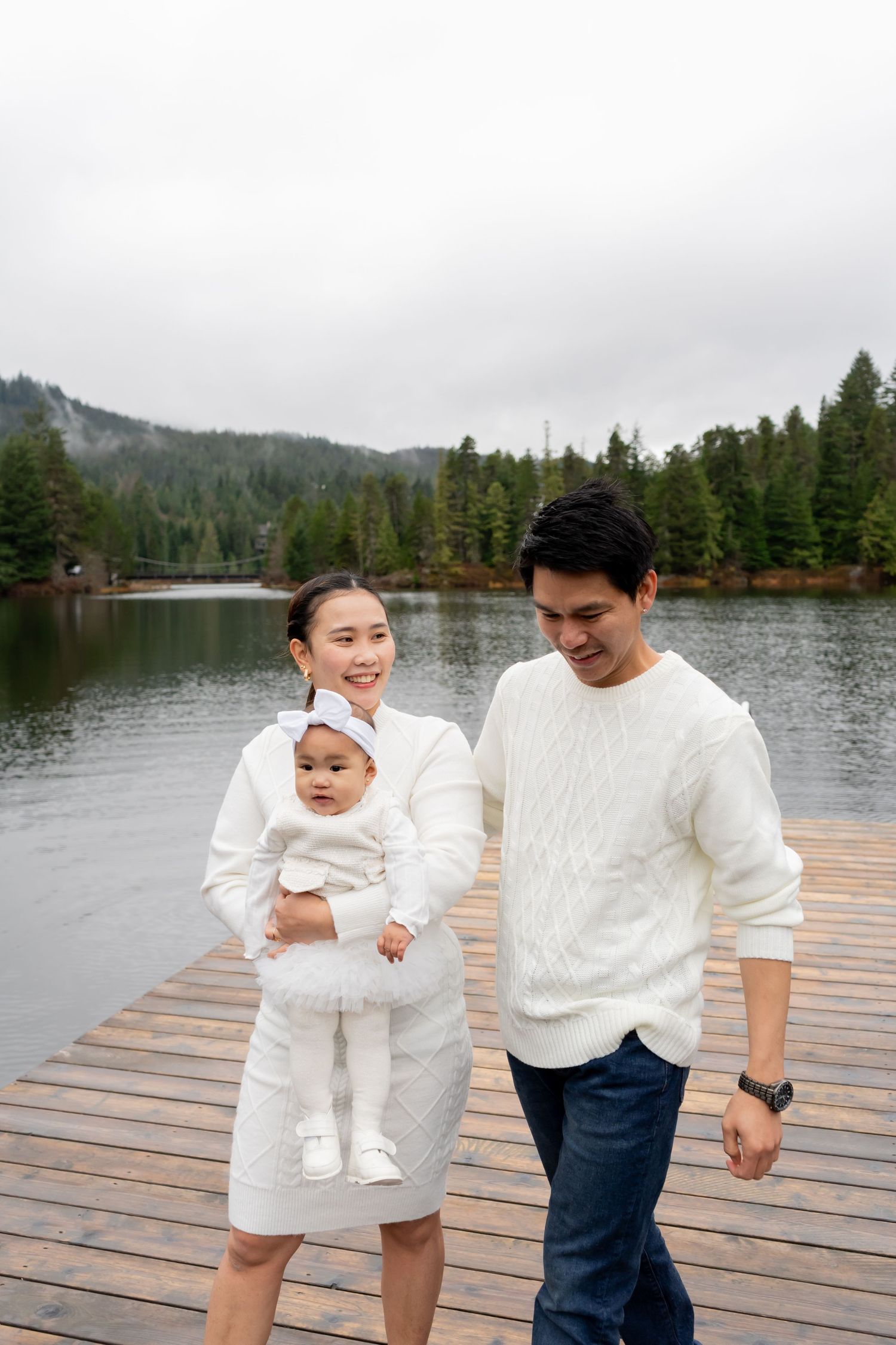 A happy family with a baby dressed in white posing on a wooden dock by a misty mountain lake.