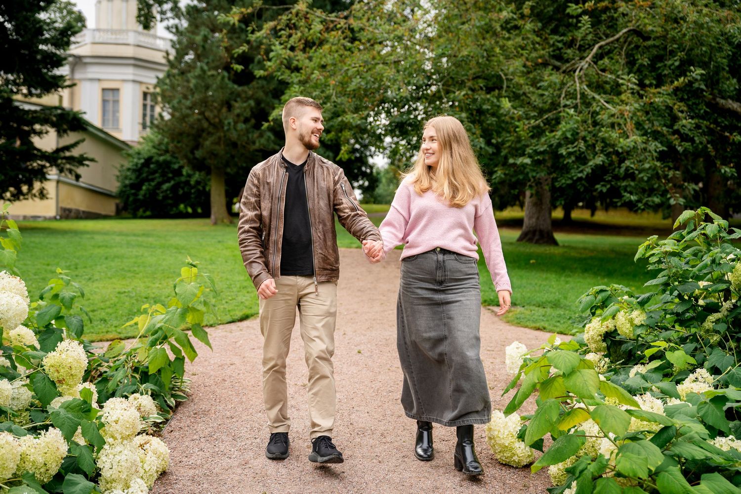 A couple walks hand in hand on a garden path surrounded by hydrangeas and lush greenery near a historic building.