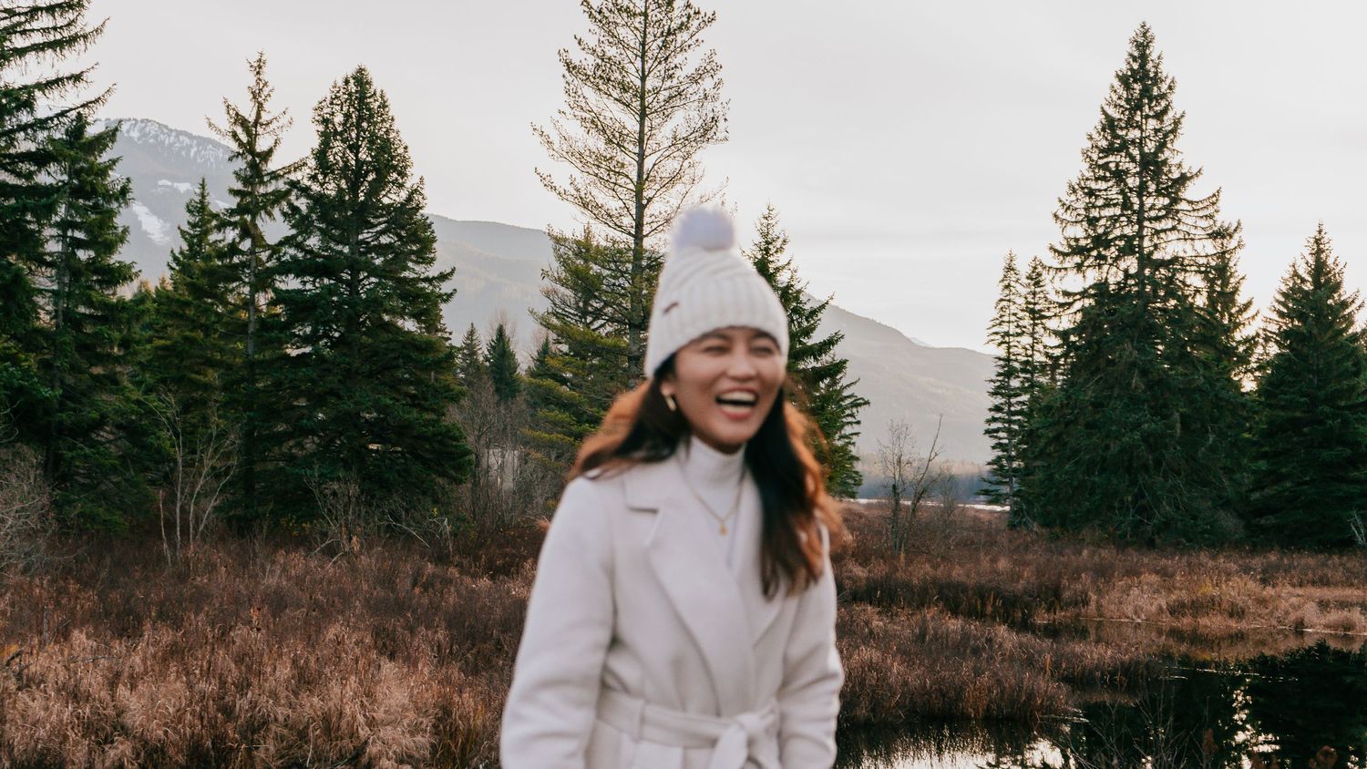 A happy woman in a white coat and pom-pom hat laughs outdoors near pine trees and mountains.