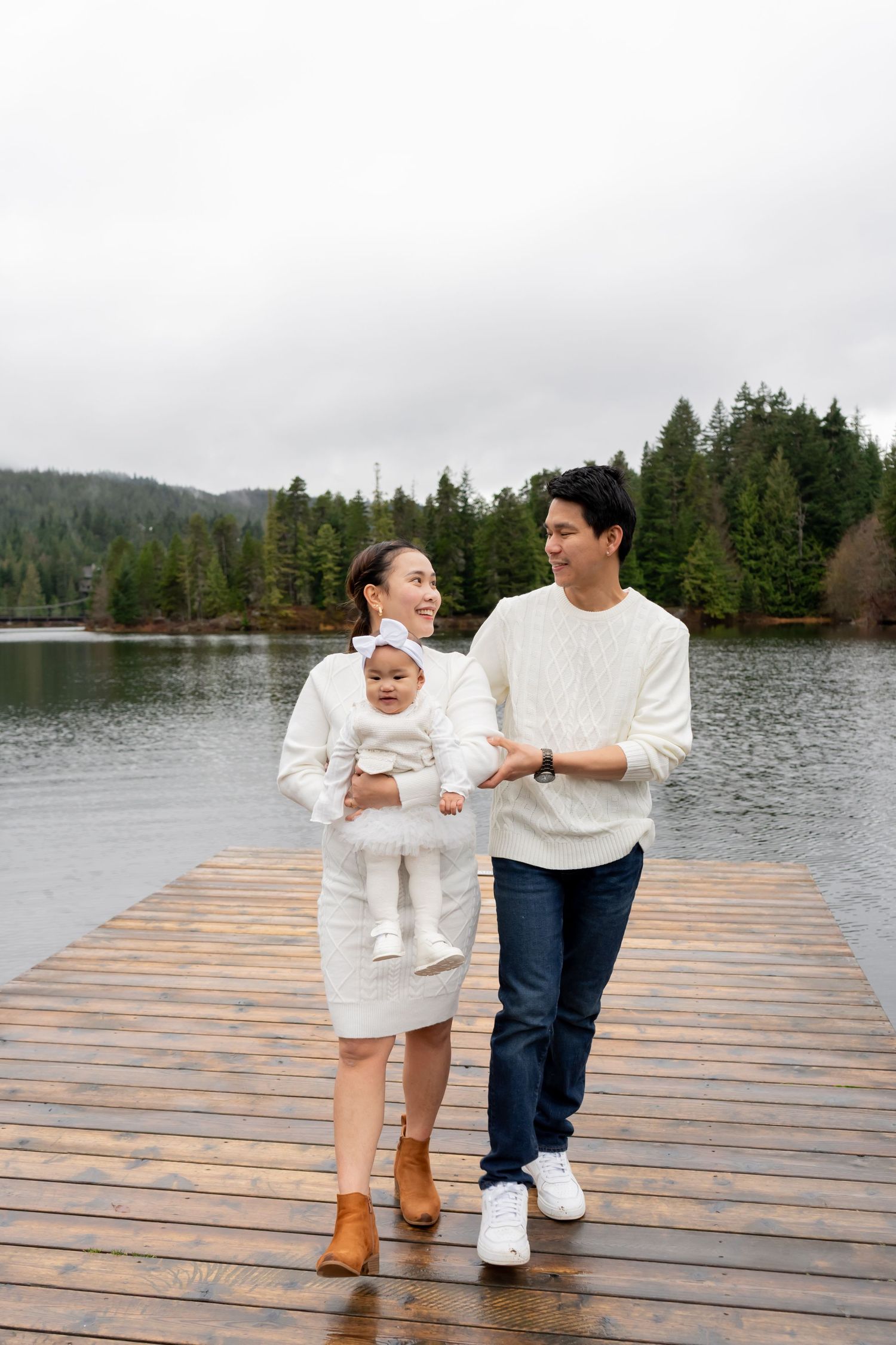 Young family in white outfits walking on a wooden dock by a lake surrounded by evergreen trees.