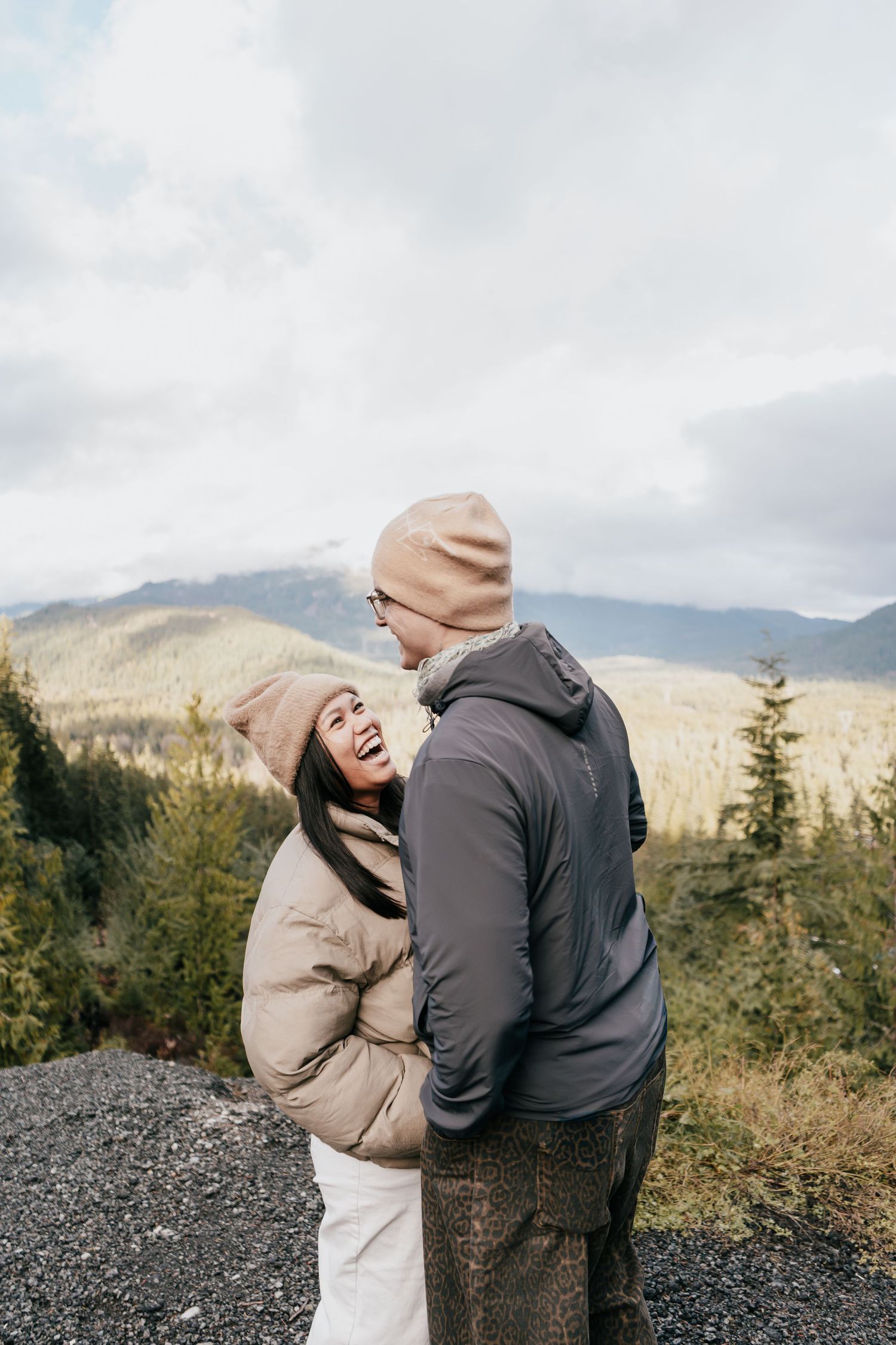 A joyful couple laughing and embracing on a rocky overlook with scenic mountain views behind them.