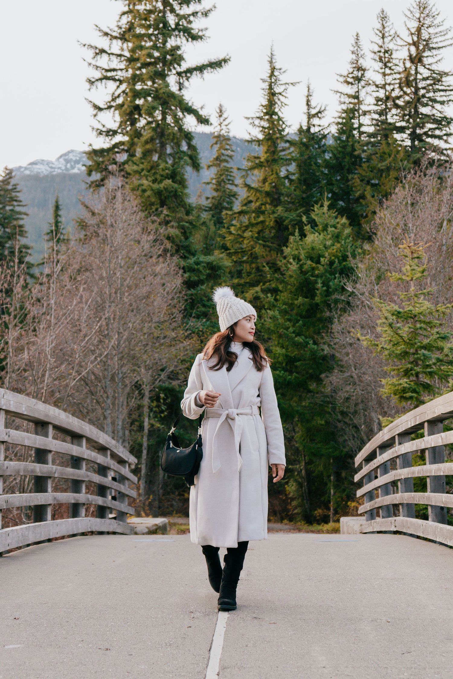 Stylish young woman in a white coat and knit hat stands on a wooden bridge surrounded by evergreen trees.