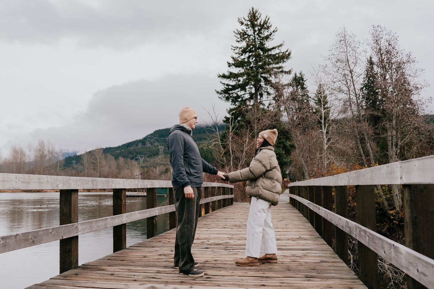 Couple holding hands on a wooden bridge overlooking a misty lake surrounded by trees.