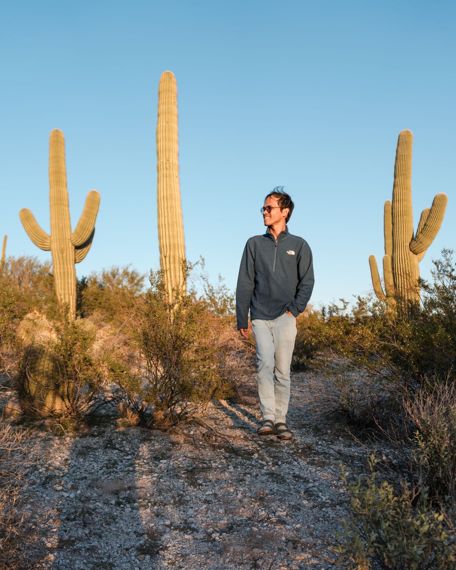 A man in a North Face jacket stands on a desert trail surrounded by tall saguaro cacti at sunset.