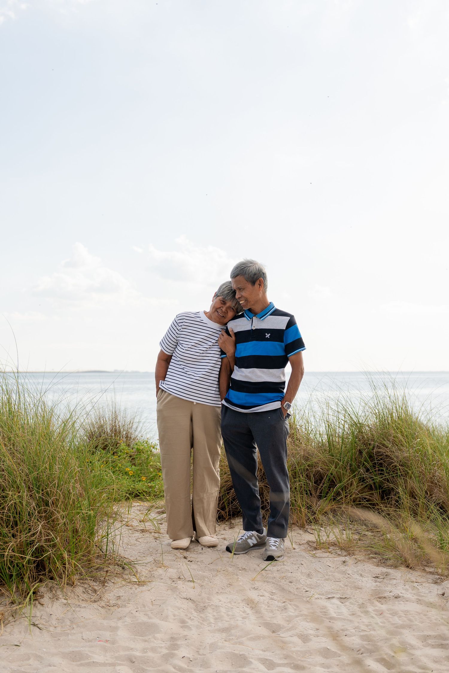 An older couple shares a tender moment on a sandy beach path with ocean views and coastal grass.
