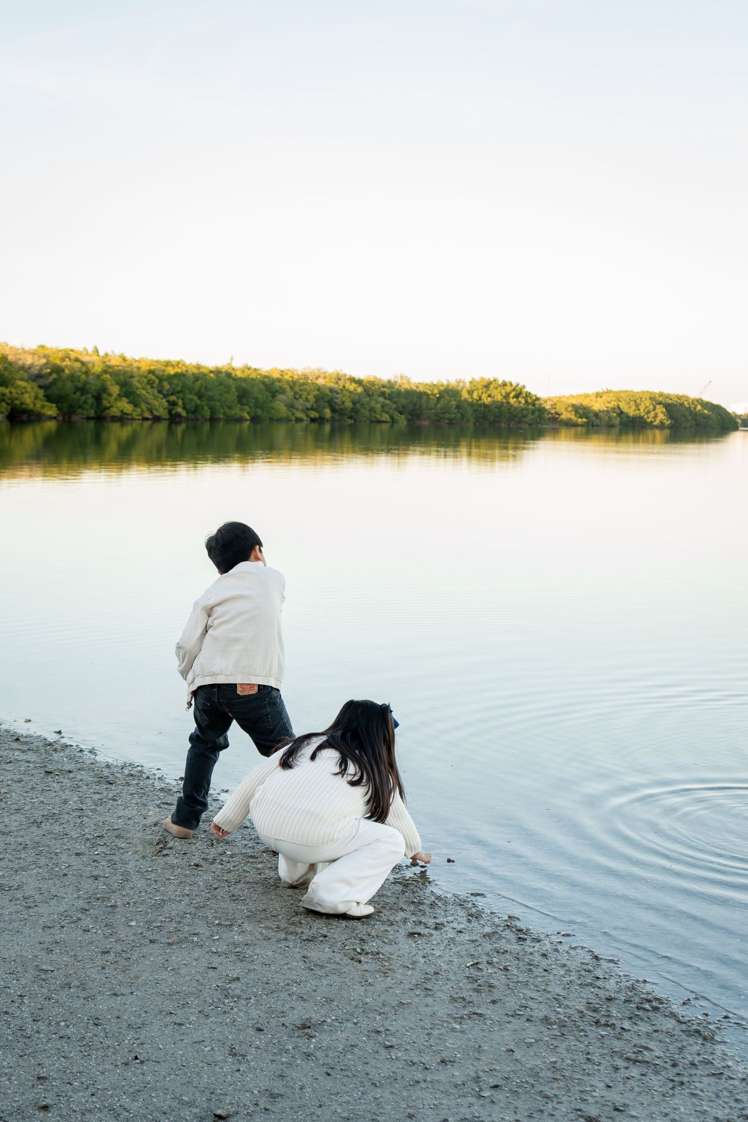 Two children crouch and play at the water's edge on a peaceful shoreline surrounded by trees.
