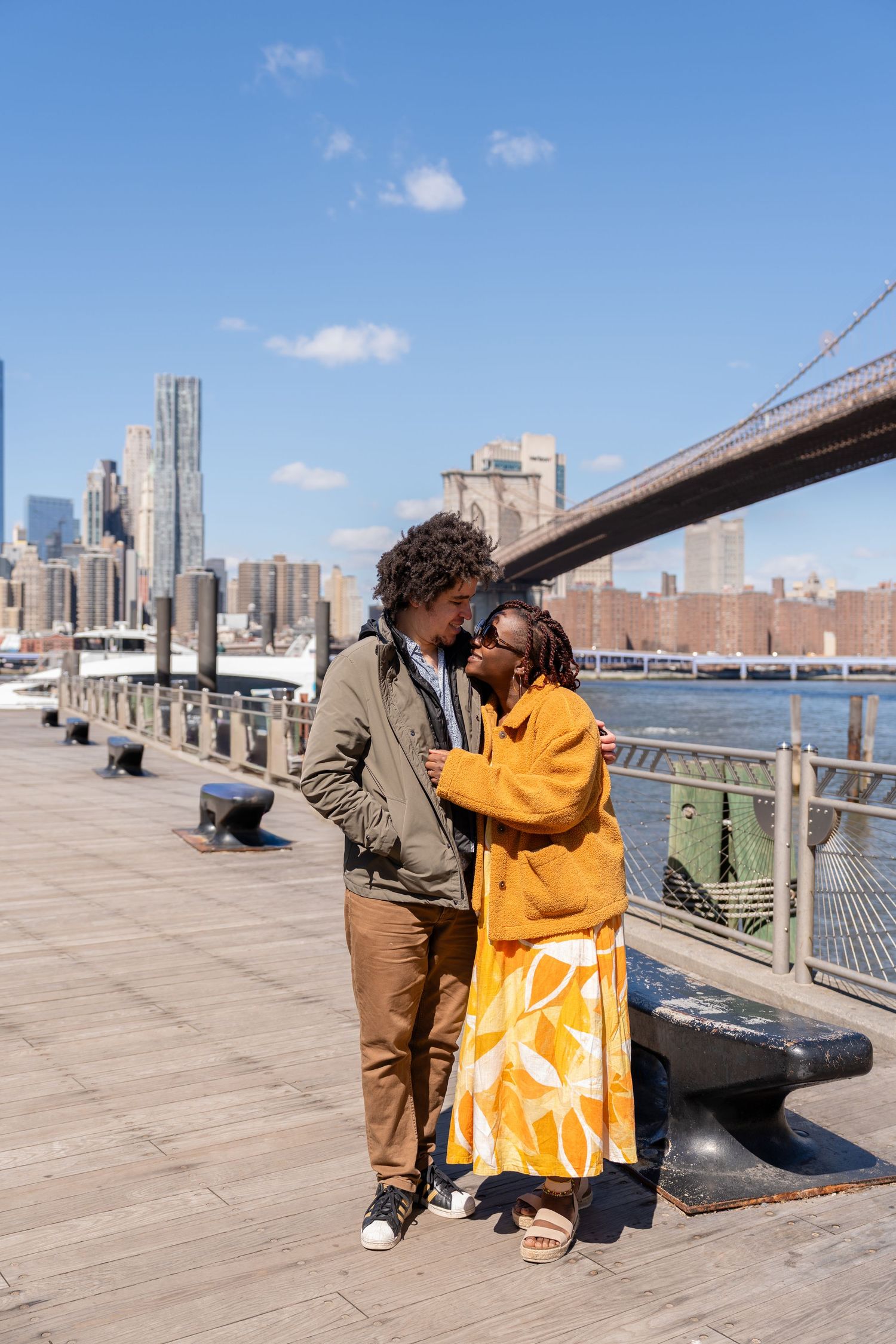 A couple shares a romantic moment on a Brooklyn waterfront boardwalk with the Brooklyn Bridge and NYC skyline behind them.