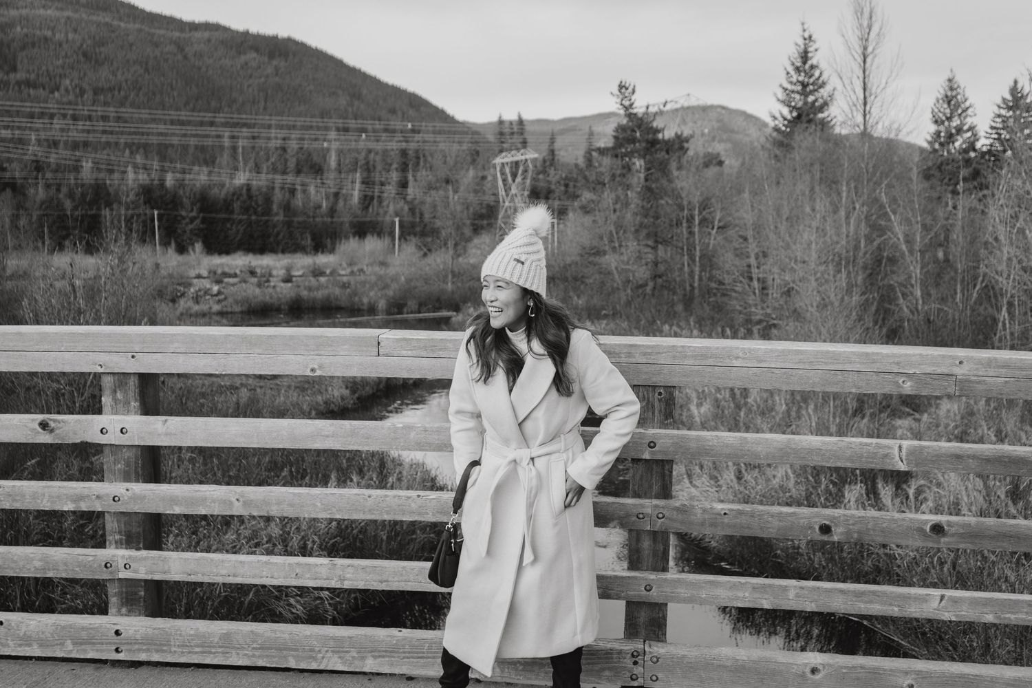 Woman in white coat and knit pom hat poses on wooden bridge with mountain backdrop.