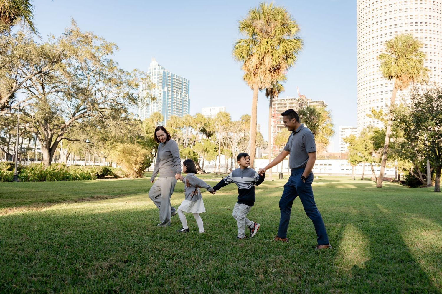 Family of four holding hands and walking in a sunny urban park with palm trees and city skyline.