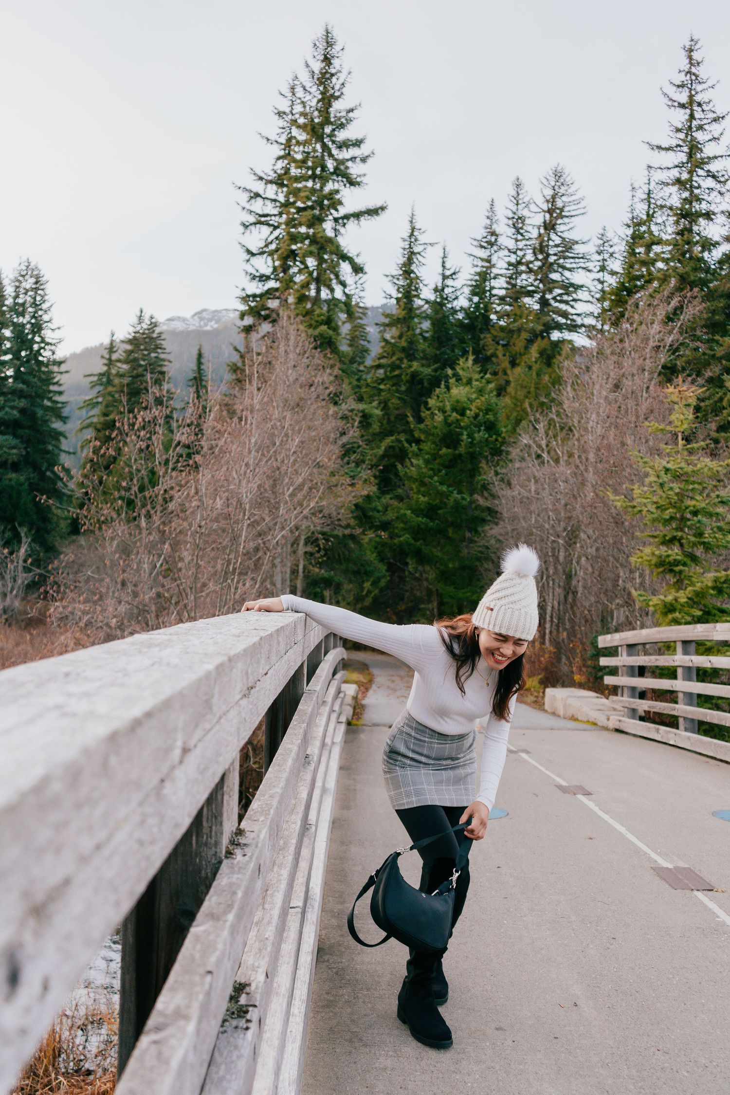 Woman in winter outfit leans on wooden bridge railing surrounded by tall evergreen trees.