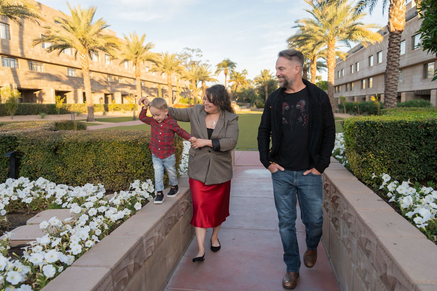 Family of three walking along a flower-lined path at a resort with palm trees and buildings.