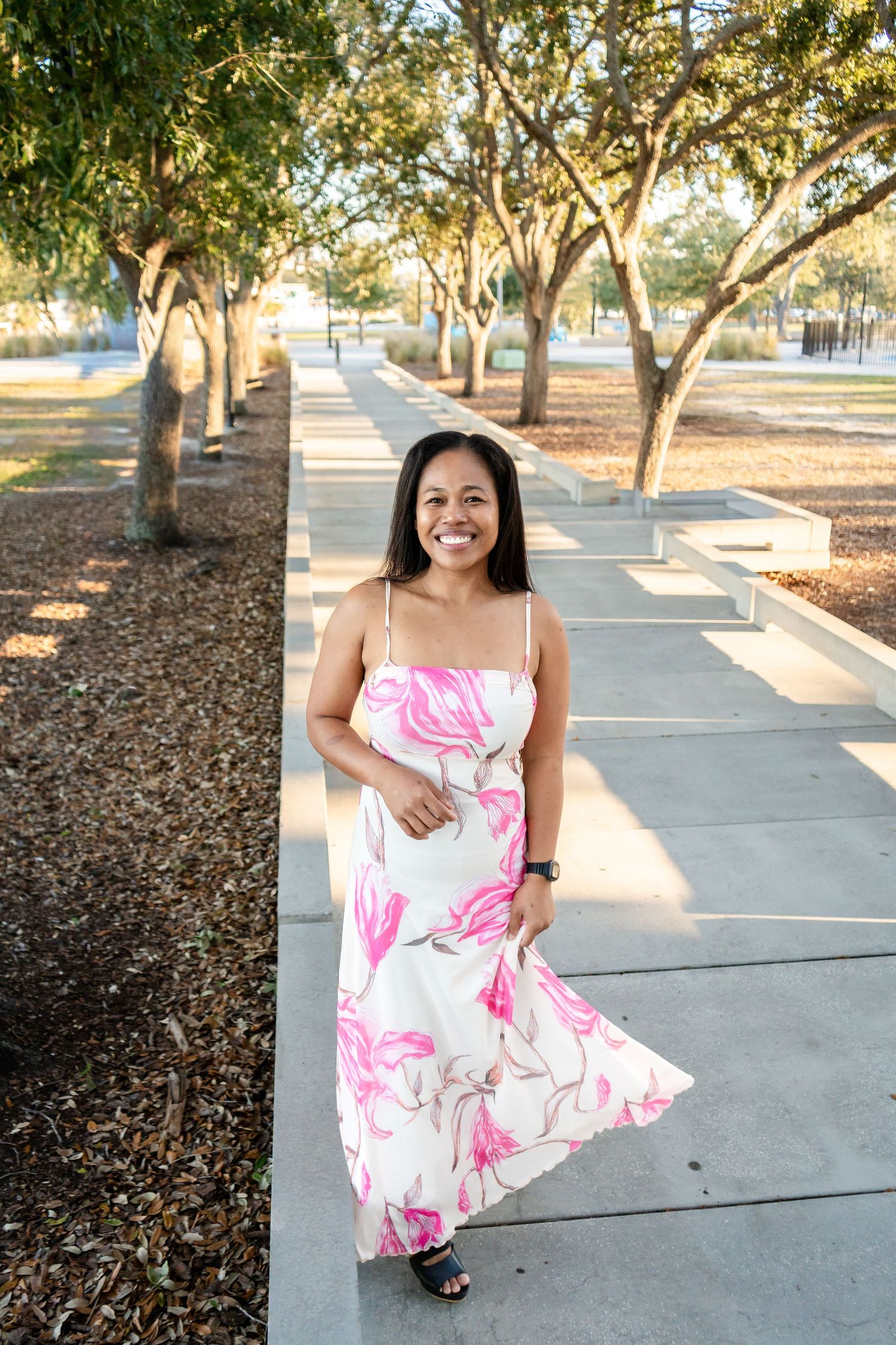 Smiling woman in white and pink floral maxi dress walking along a tree-lined outdoor path.