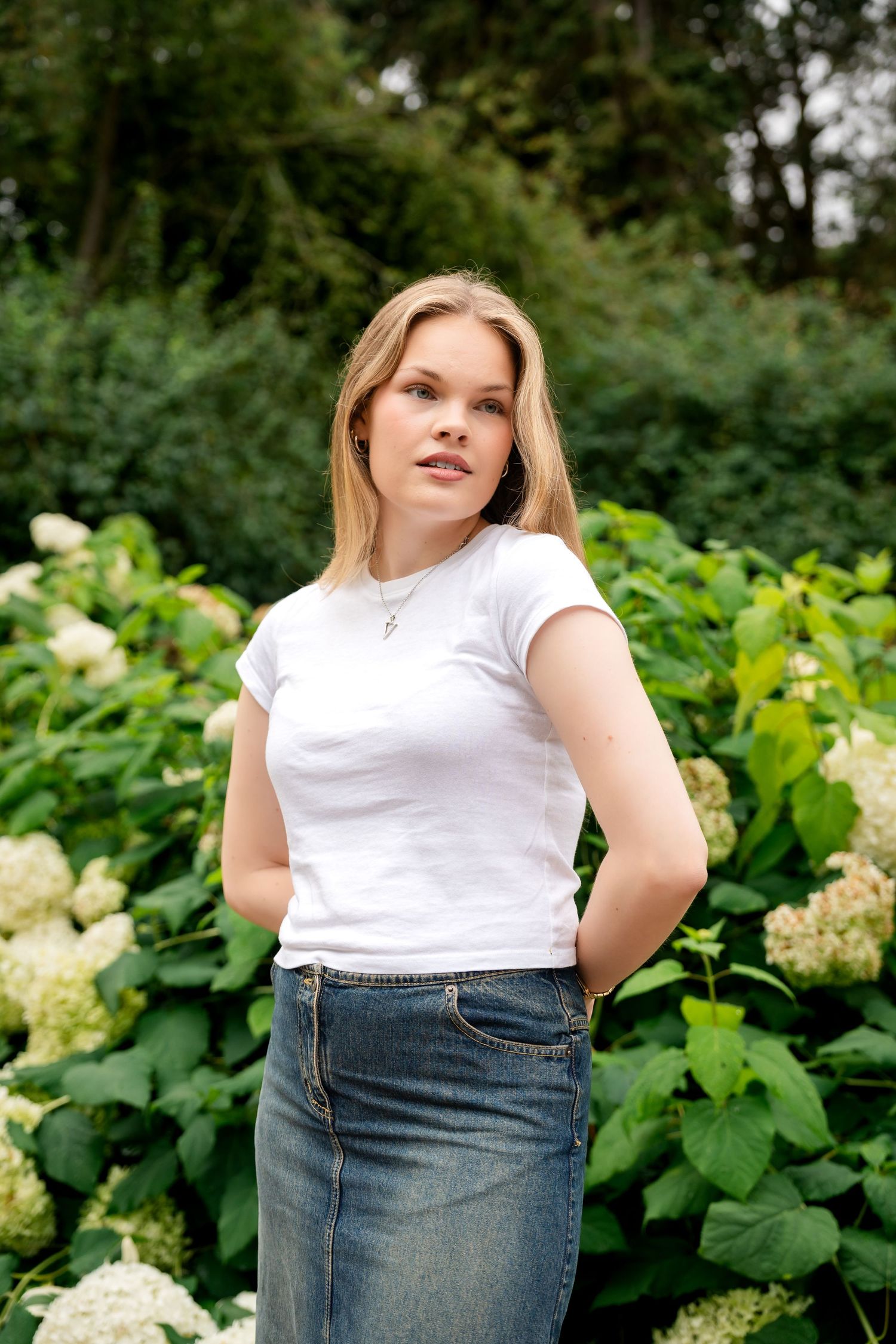 Young blonde woman in white t-shirt and denim skirt posing outdoors among lush green hydrangea bushes.