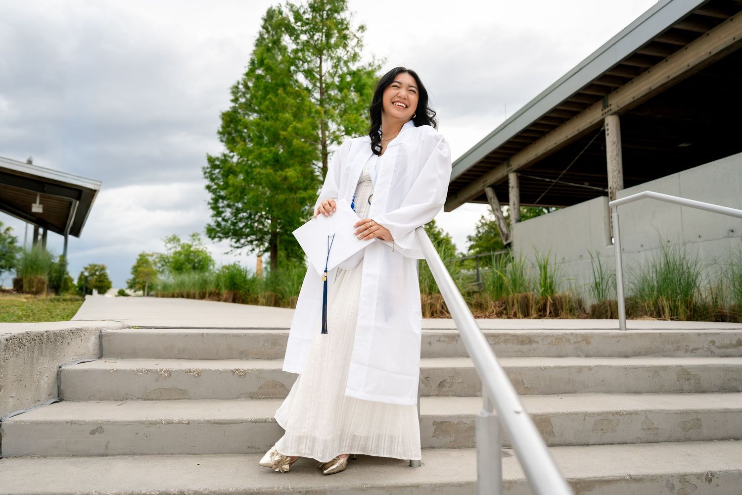 A young woman in white graduation robes poses on outdoor steps holding her diploma with a big smile.